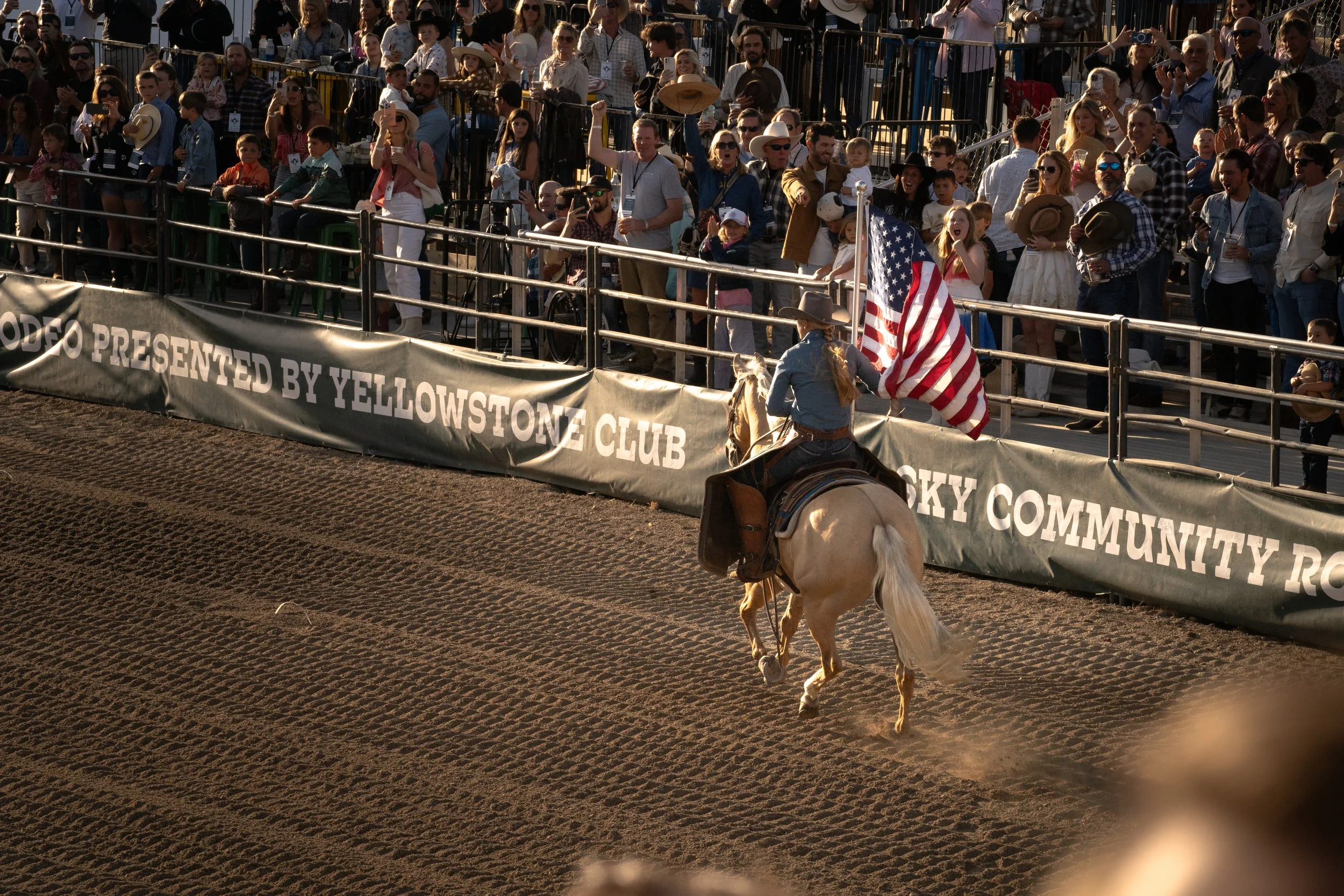 A rider on a white horse holding an American flag at a rodeo. Spectators are watching from the stands, some taking photos, under late afternoon sunlight.