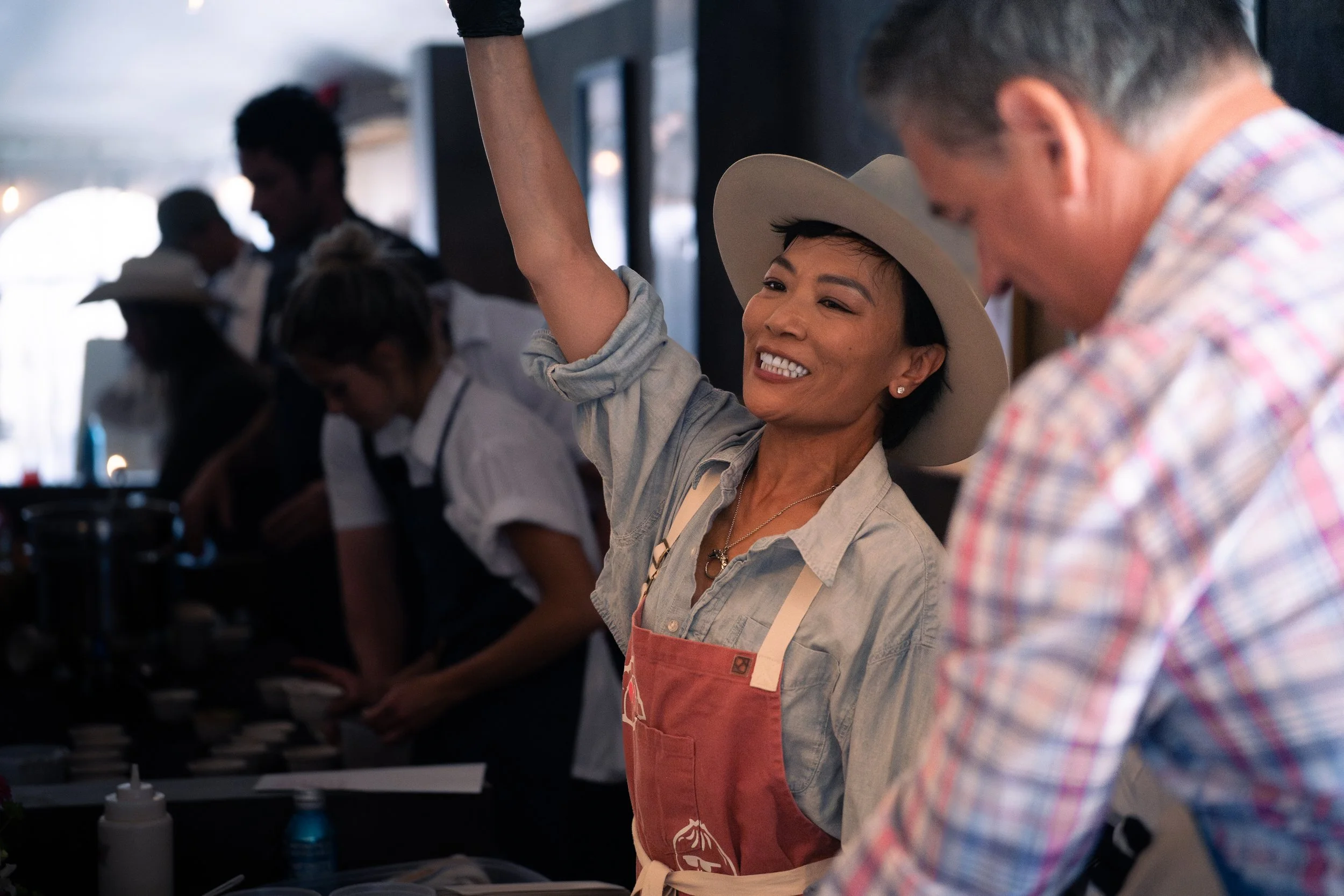 A smiling woman with short dark hair, wearing a beige wide-brimmed hat, a light gray shirt, and a pink apron, raising her right arm. She is in a busy indoor setting with people working behind her, likely in a kitchen or café.