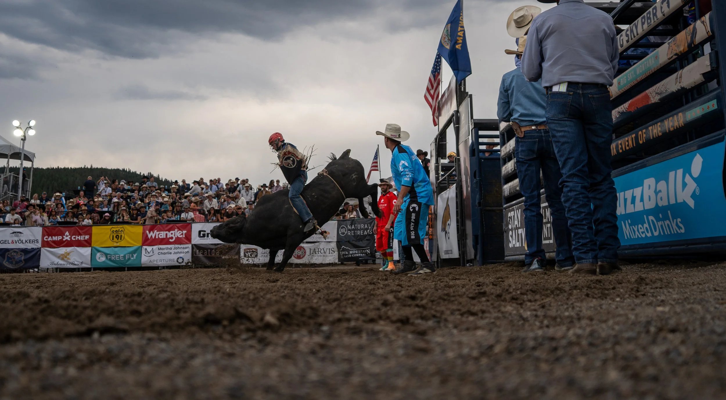 Rodeo event with a bull rider falling off a bucking bull while helpers and spectators watch under cloudy skies.