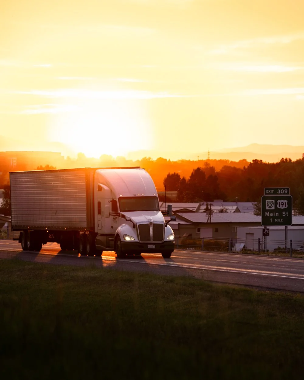 A semi-truck driving on a highway during sunset with orange sky and some clouds, houses, and road signs nearby.