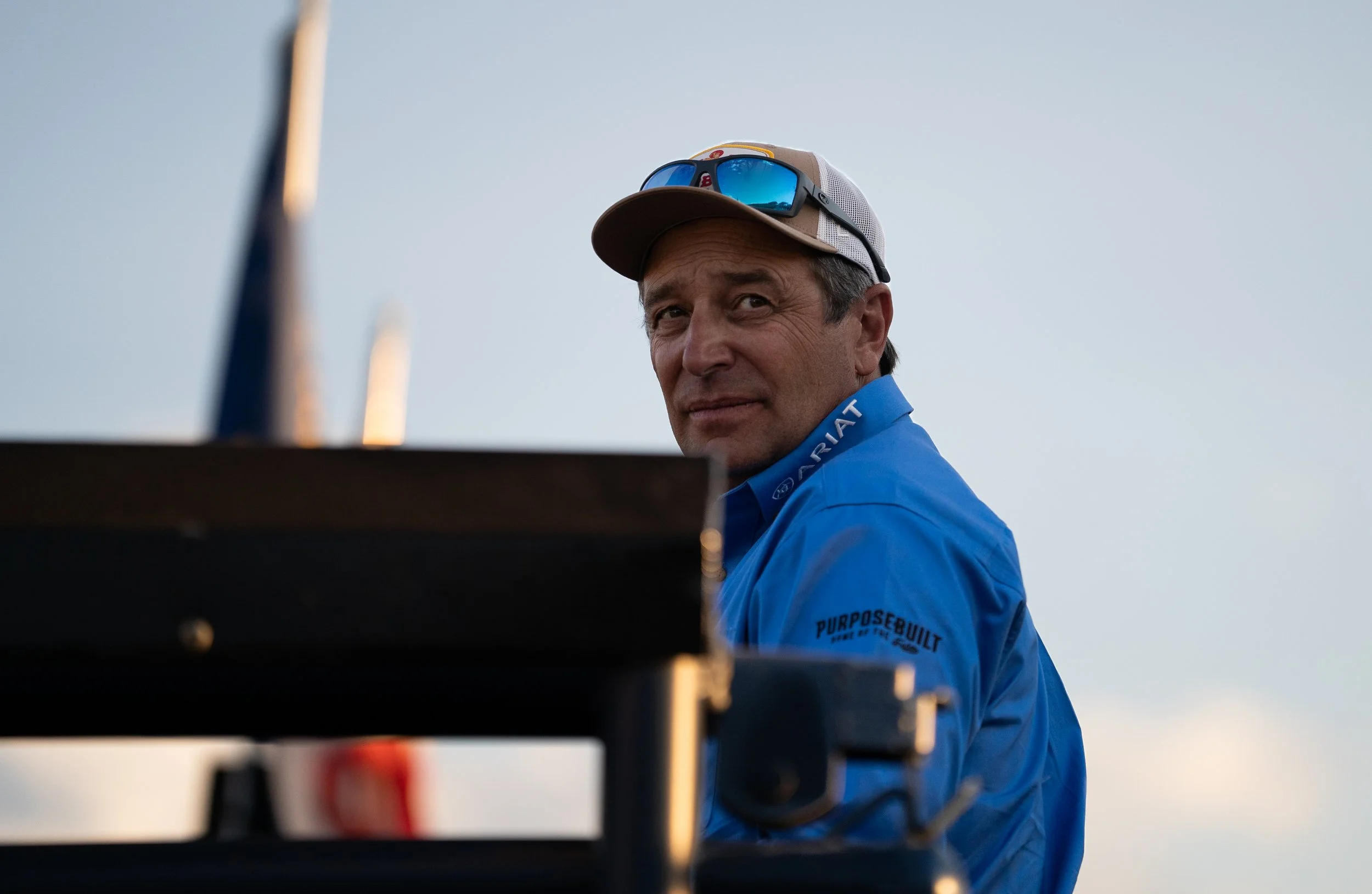 A man wearing a blue Patagonia jacket, sunglasses on his hat, and a baseball cap, looking over a piece of equipment against a clear sky.