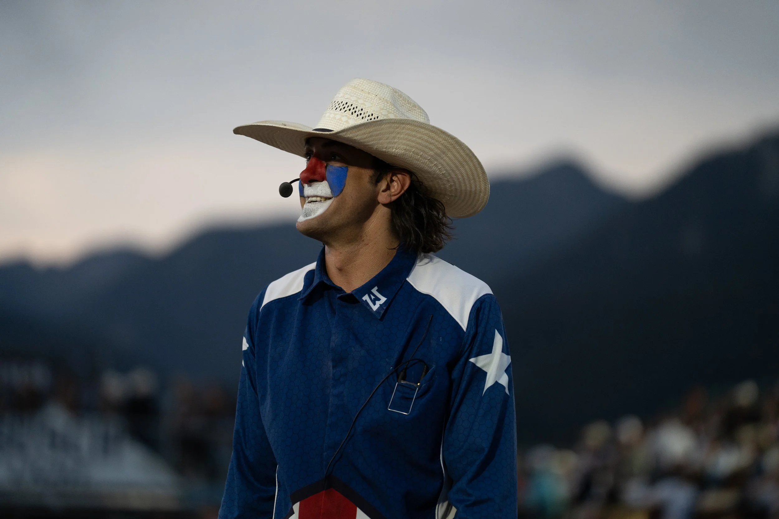 A man dressed as a clown wearing a cowboy hat and blue racing suit with white and red accents, standing outdoors at dusk or dawn, with mountain silhouettes in the background.