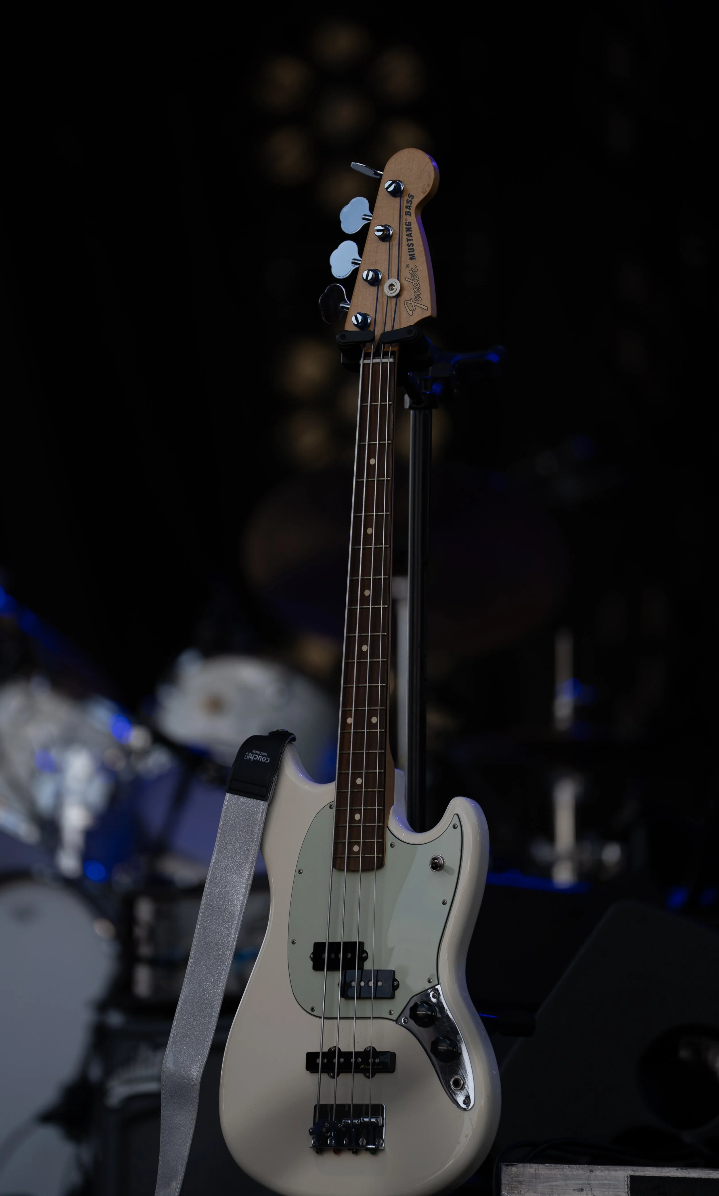 An electric bass guitar on a stand with a strap, in a dimly lit stage setting, with drums blurred in the background.