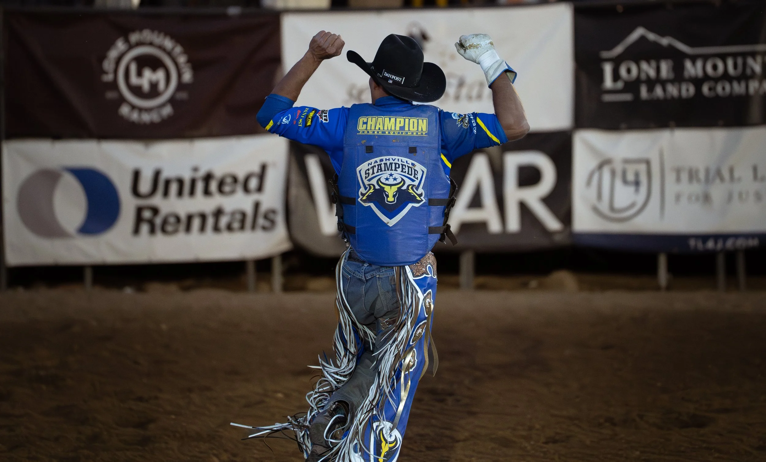 A person in a blue Nashville Stampede jacket and matching pants with fringe, wearing a black cowboy hat and gloves, celebrates with raised arms on a dirt arena. Banners with sponsor logos are in the background.