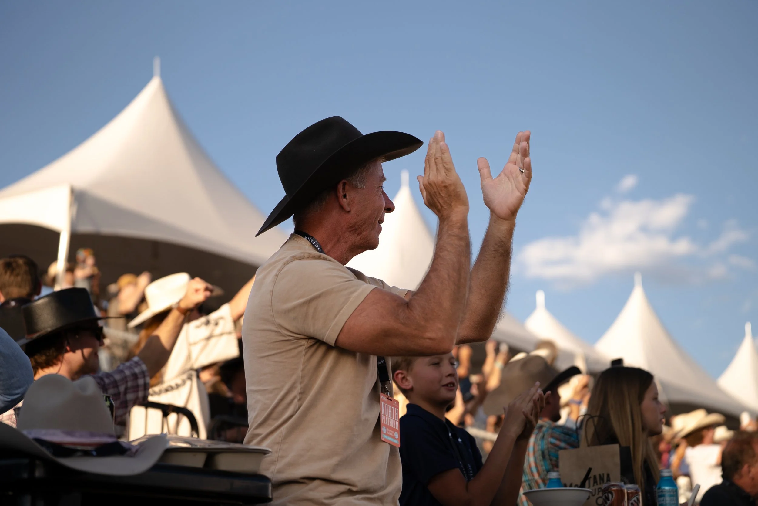 A crowd of people, including an older man in a beige shirt and black wide-brimmed hat, clapping and enjoying an outdoor event under a clear blue sky with white tents in the background.