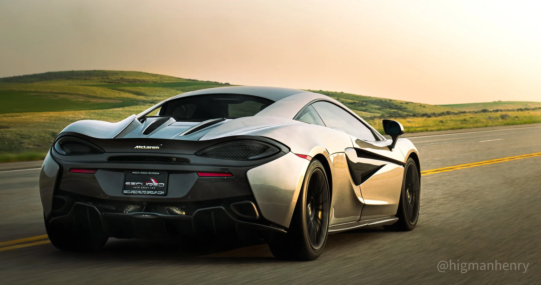A silver McLaren sports car driving on an open road with green hills in the background at sunset.