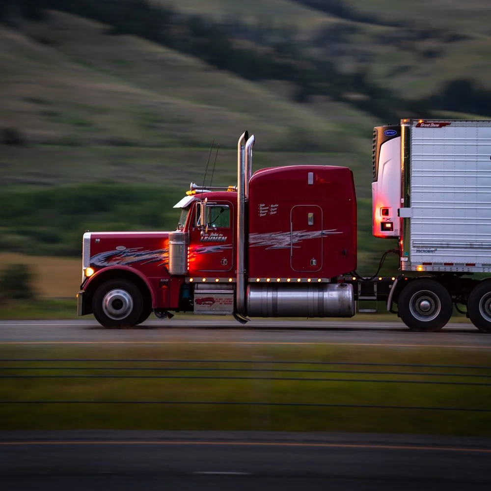 A red semi-truck driving on a highway with a blurred green hillside in the background at dusk.