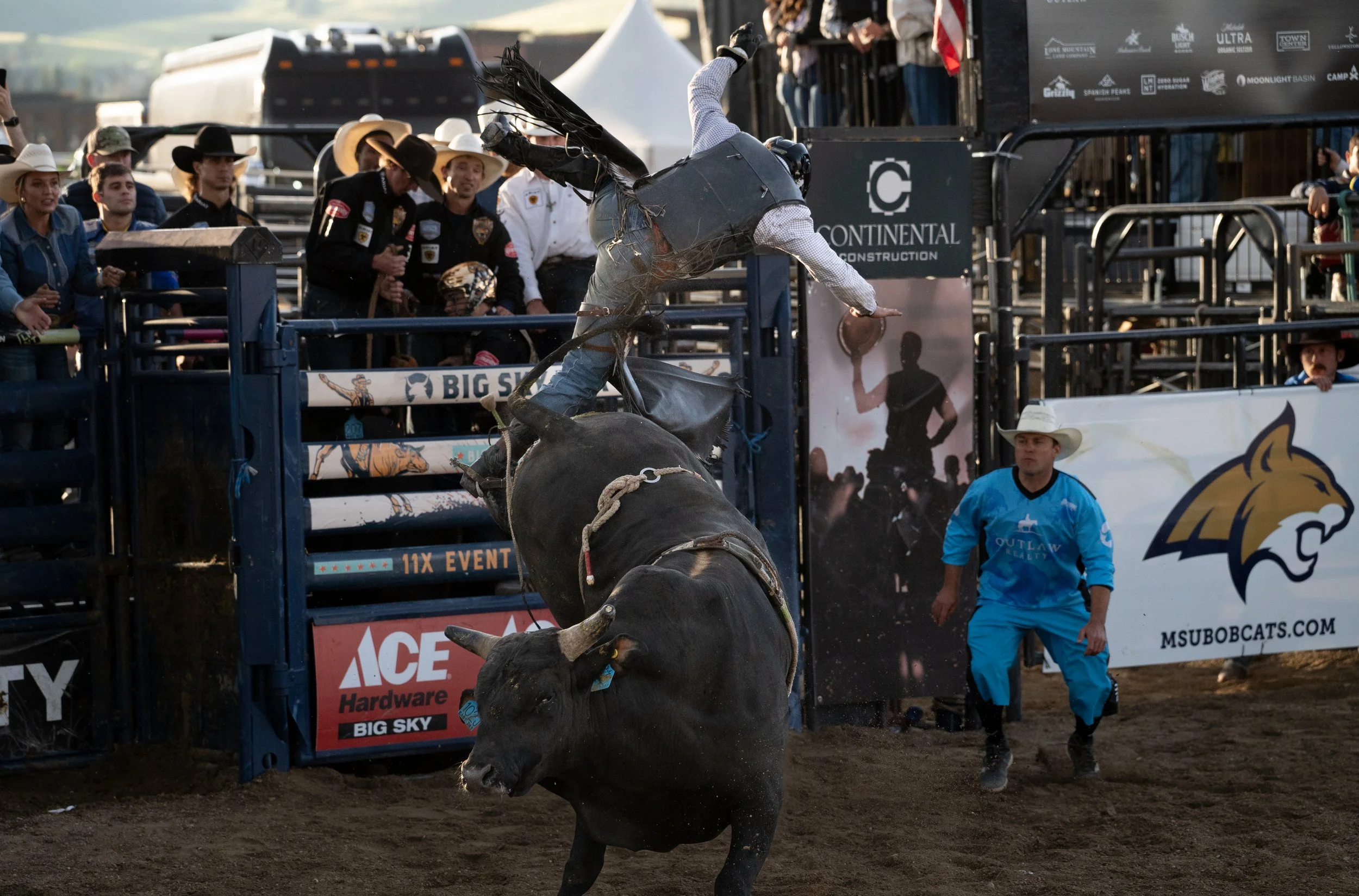 A rodeo event where a cowboy loses his hat and is thrown off a bucking bull. Spectators watch from behind the fence, and a rodeo clown in blue stands nearby.