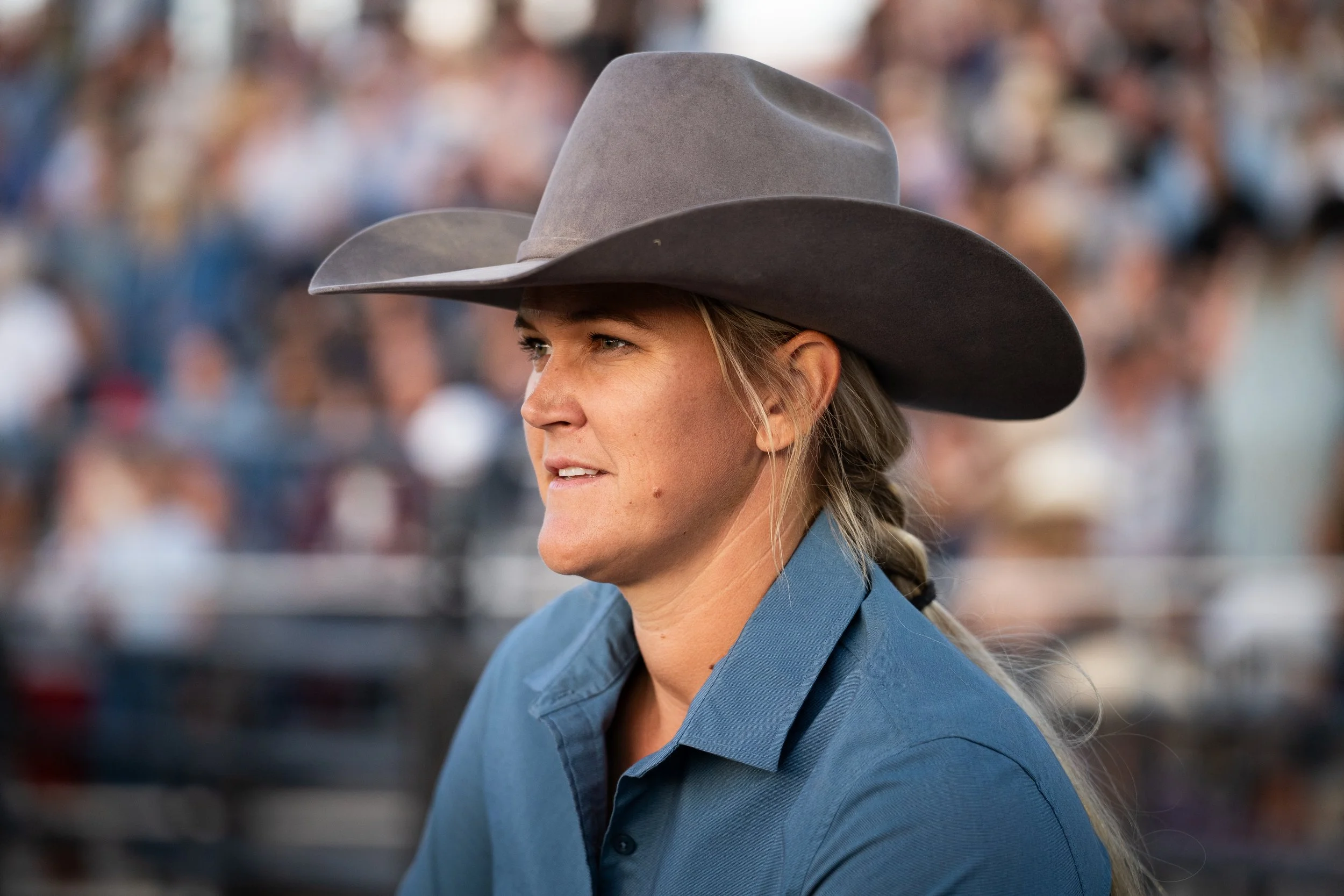 A woman wearing a cowboy hat and a blue shirt, with a braided hairstyle, is outdoors with a crowd in the background.