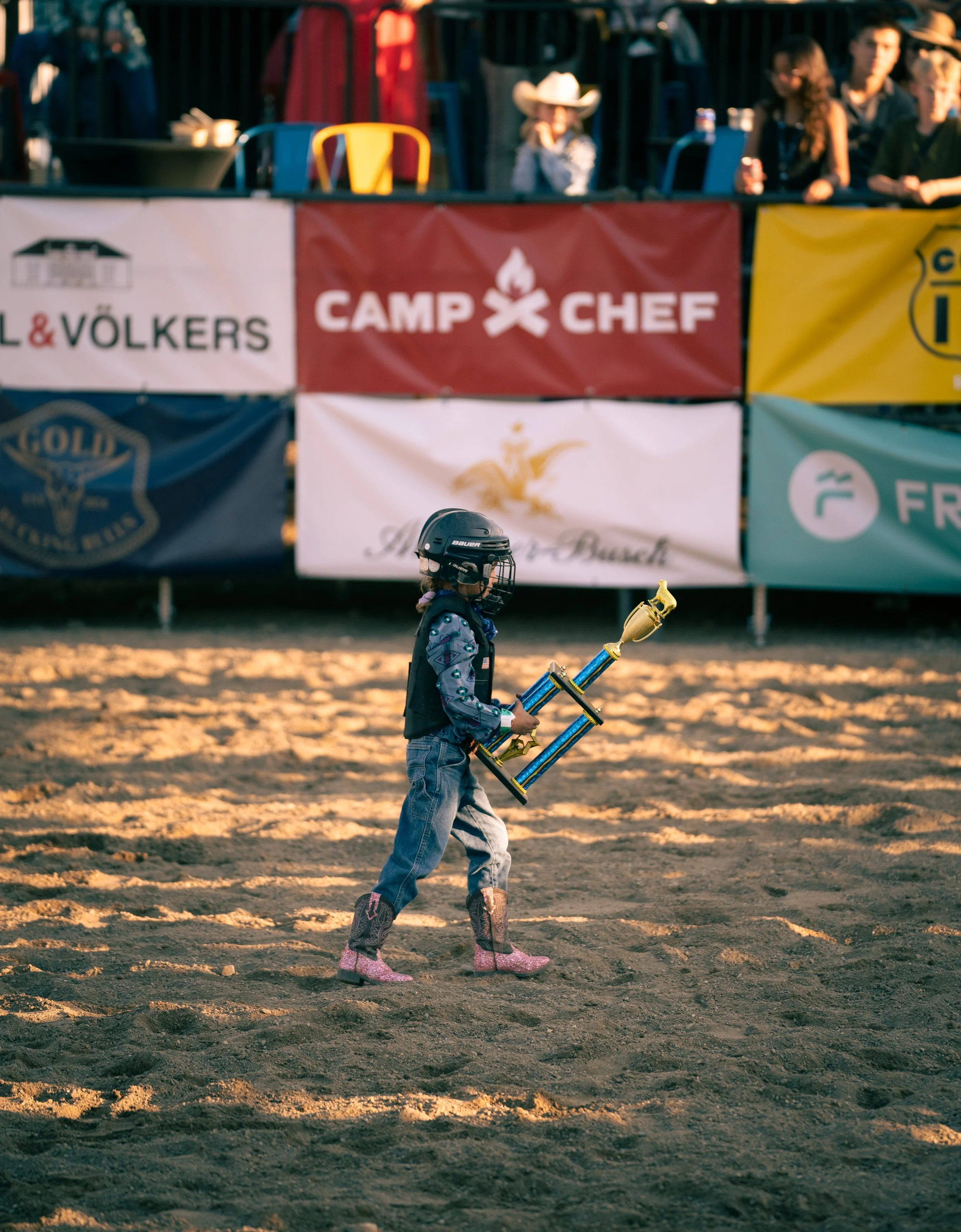 A young girl in hockey gear, including a helmet and pink boots, walking on a dirt field while holding a large trophy with a gold cup and two blue columns. In the background, there are banners with the words "Camp Chef" and other logos, and spectators