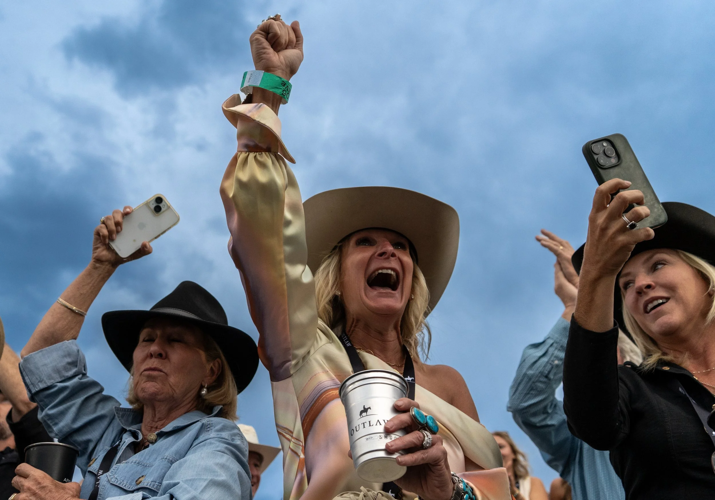 Women wearing cowboy hats and Western attire at an outdoor event, some raising their fists and holding smartphones, with a cloudy sky in the background.