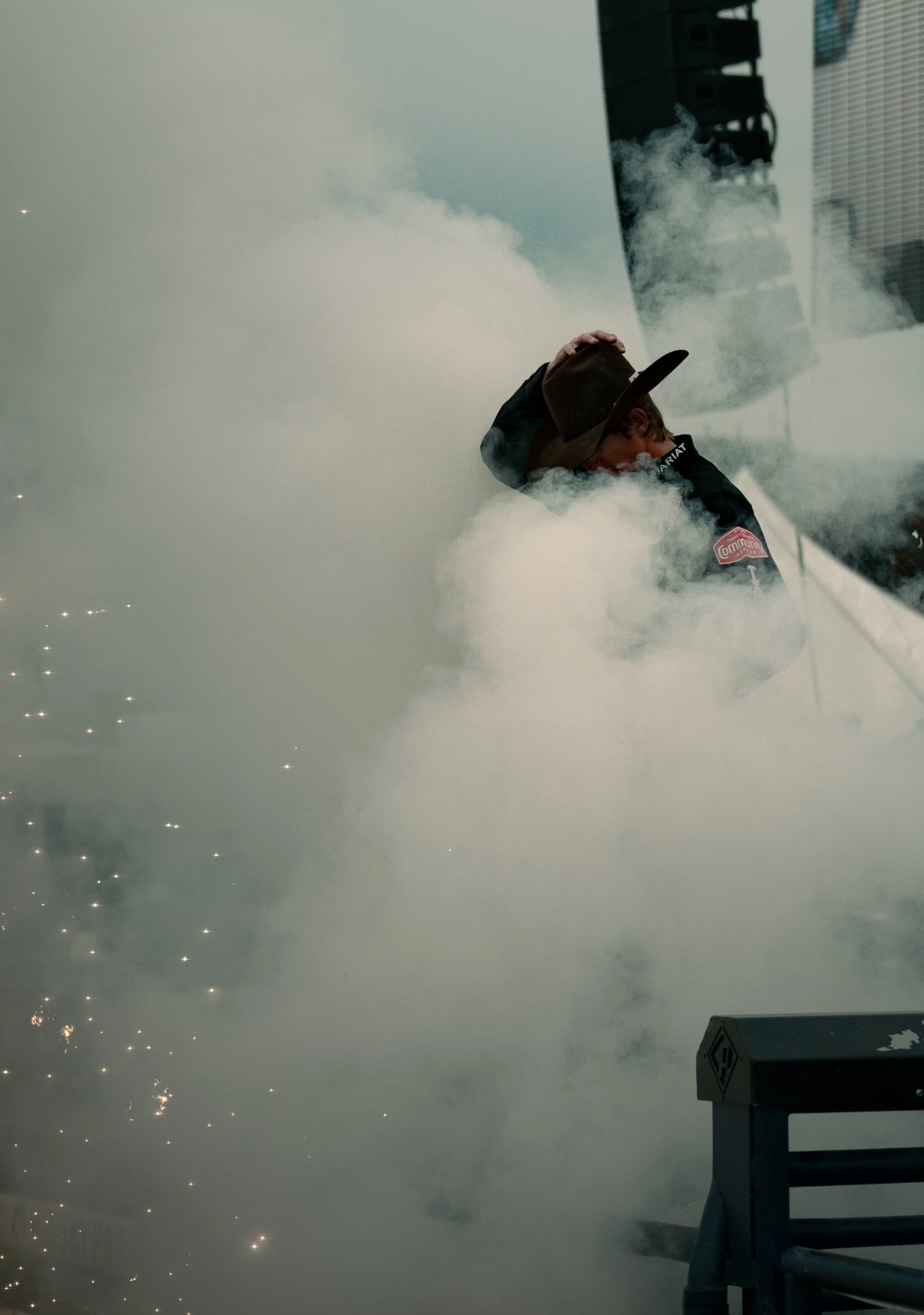 A person wearing a hat and a jacket with onlookers in the background, surrounded by thick white smoke or fog during an outdoor event.