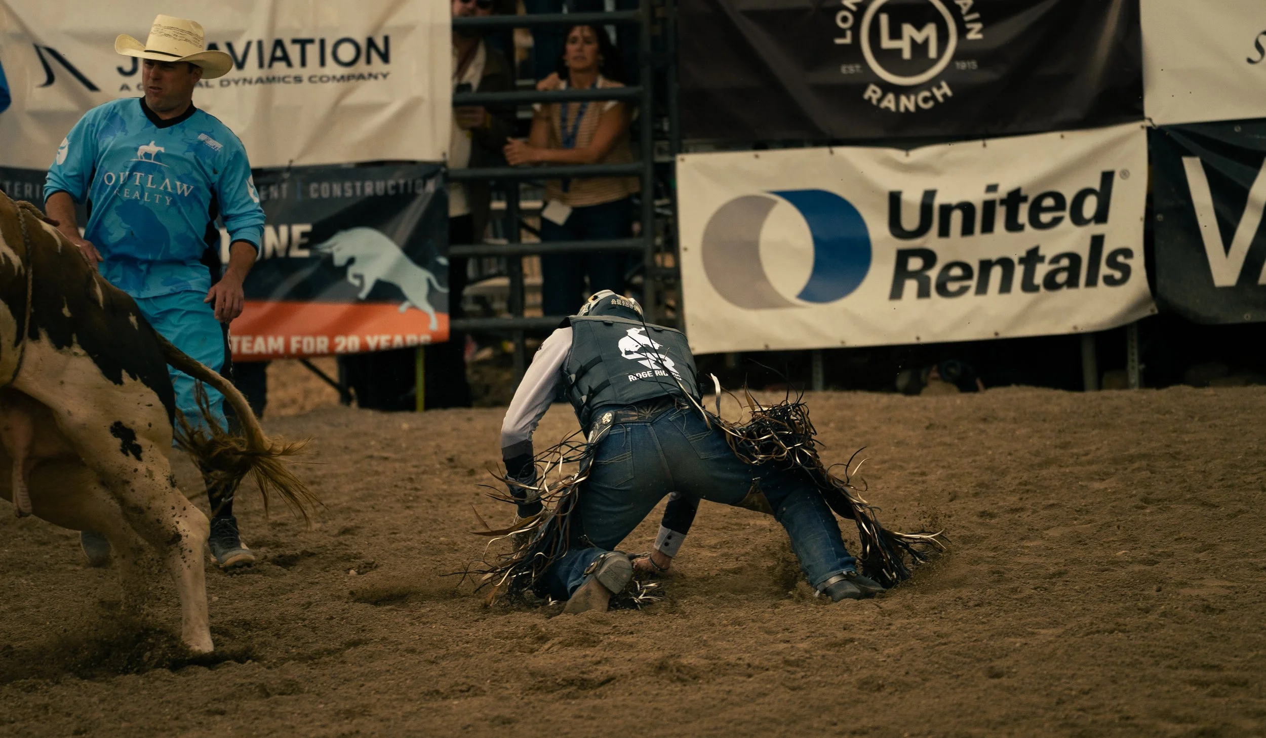 A rodeo cowboy in jeans and a vest covered in rope is falling to the dirt ground during a rodeo event, with a bull partially visible on the left side of the image. A cowboy in a light blue shirt and a cowboy in a white hat are nearby. There are banne