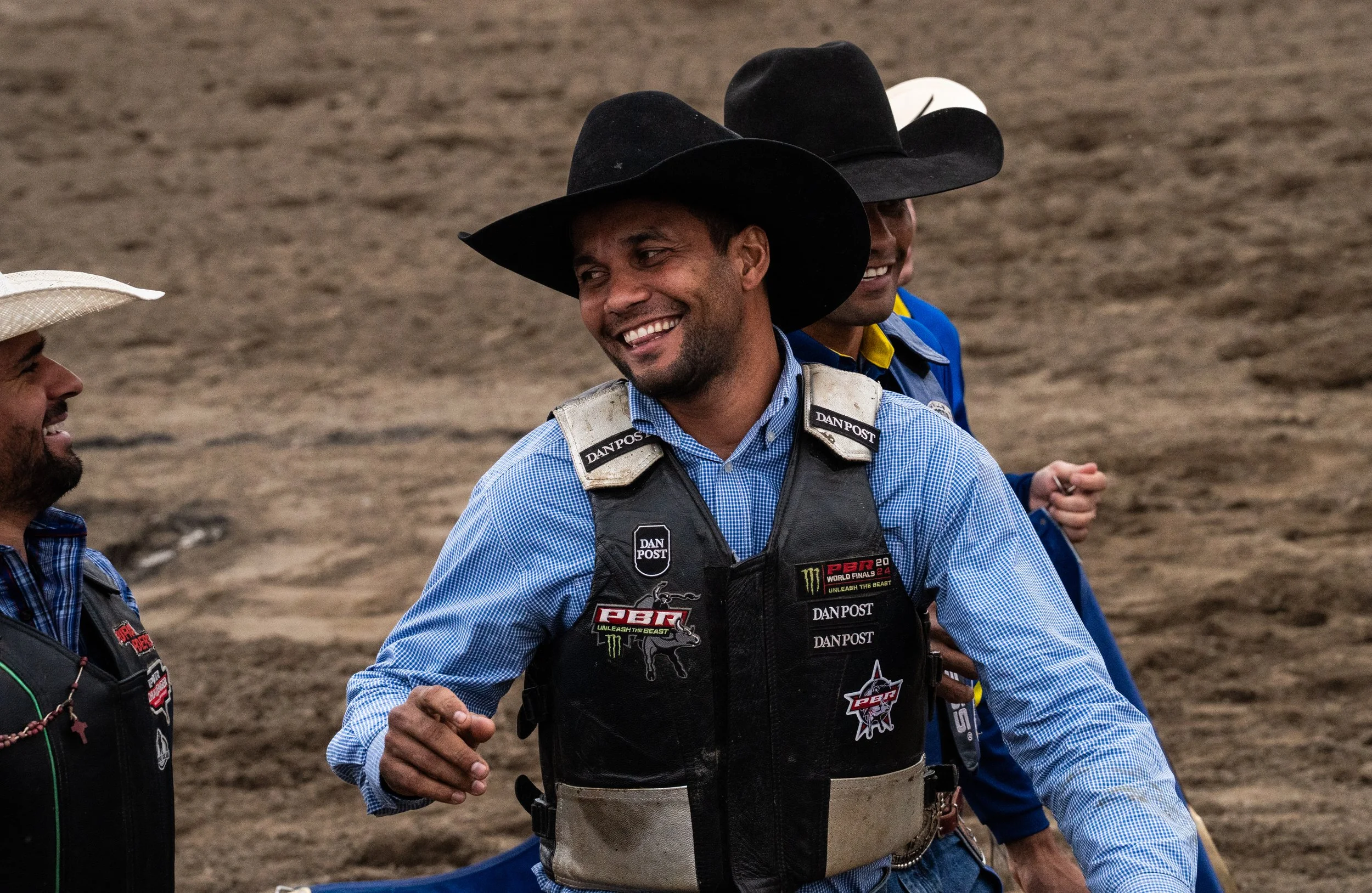 A group of men dressed in rodeo or bull riding attire, smiling and talking outdoors on dirt ground, with one man in the foreground wearing a black cowboy hat and a vest with sponsor patches.