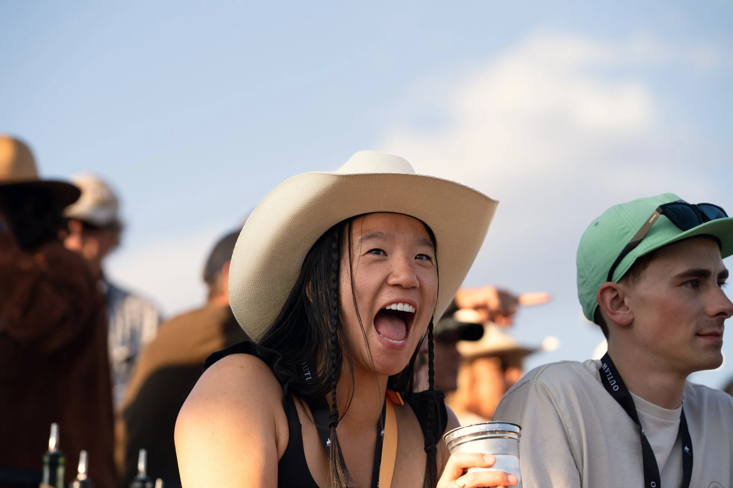 A woman wearing a large sun hat and holding a drink, smiling and appearing to shout or cheer, at an outdoor event with people in the background.