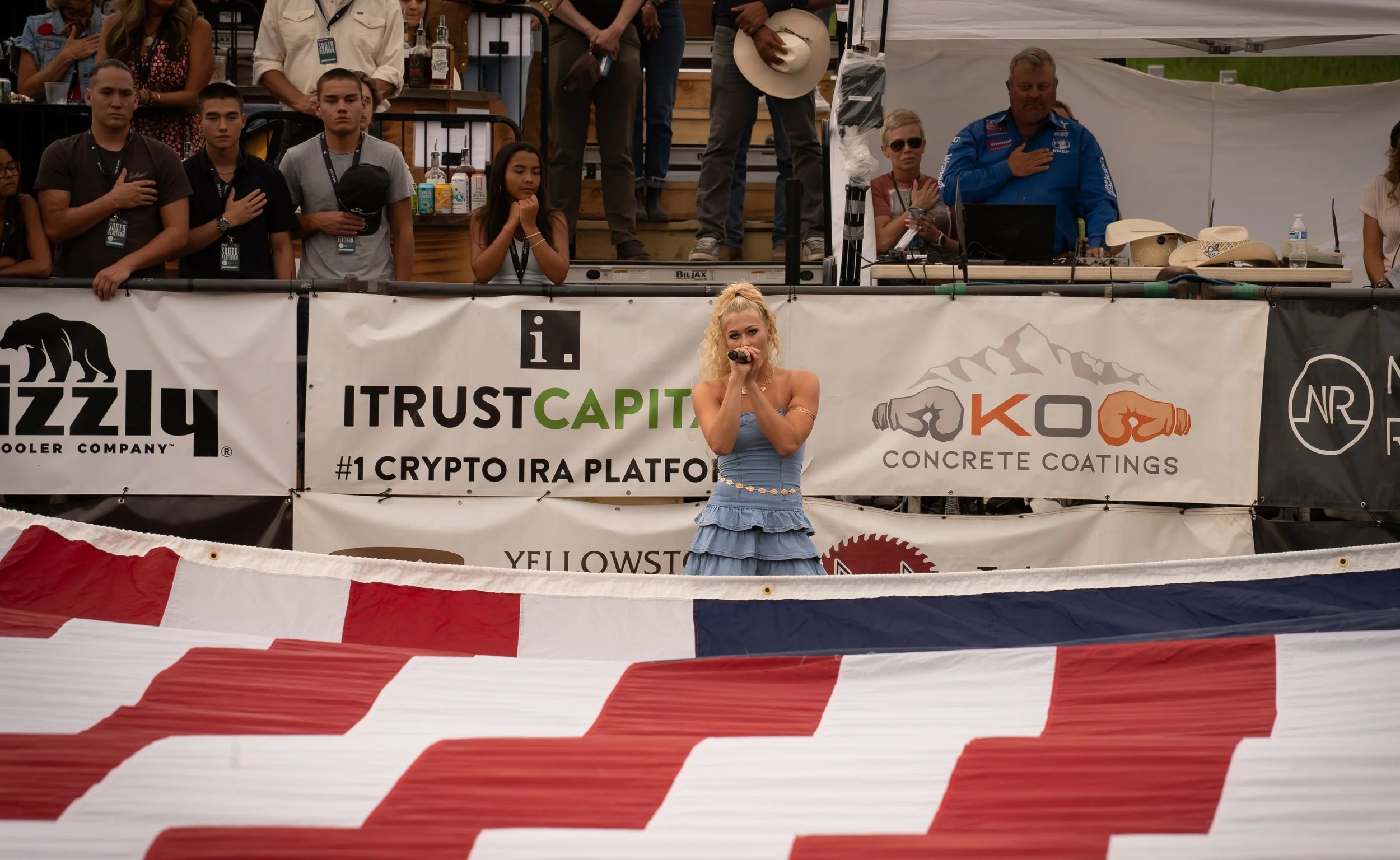 A woman singing on stage at a public event with an audience and various banners behind her.
