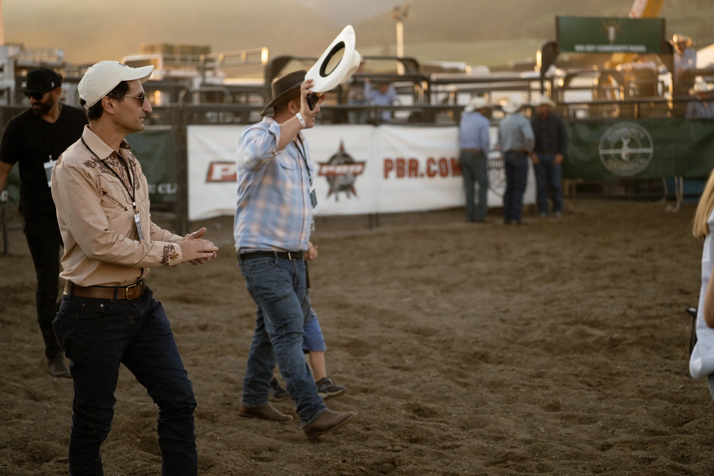 People standing and walking on dirt ground at a rodeo or outdoor event with banners and fencing in the background.