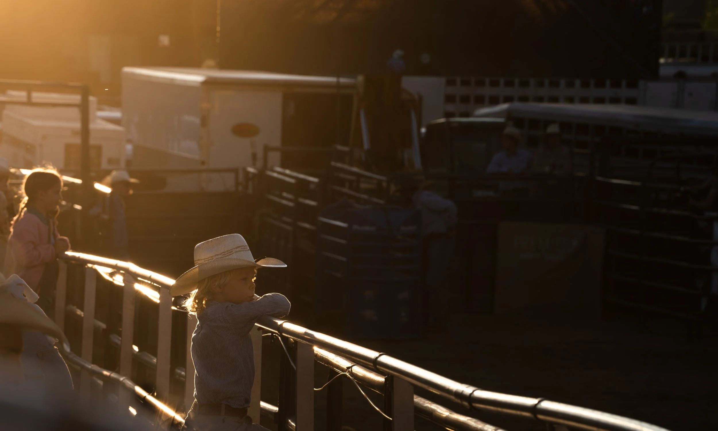 Children at a rodeo event during sunset, with one child leaning on a fence and wearing a cowboy hat, and others in the background, some wearing cowboy hats.