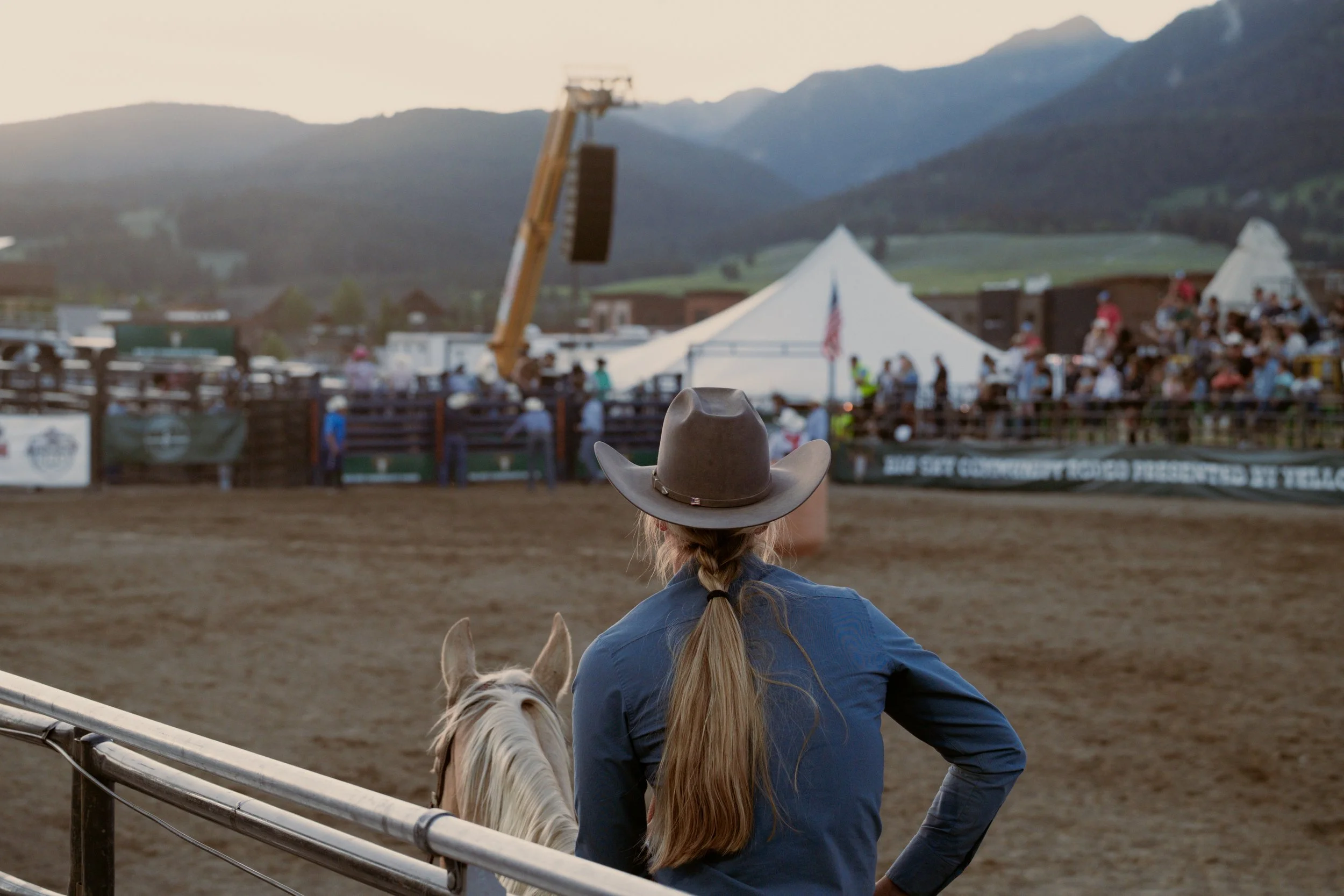 A woman with long hair in a braid wearing a cowboy hat and blue shirt, sitting on a white horse, watching what appears to be a rodeo event with a crowd, tents, and mountains in the background.