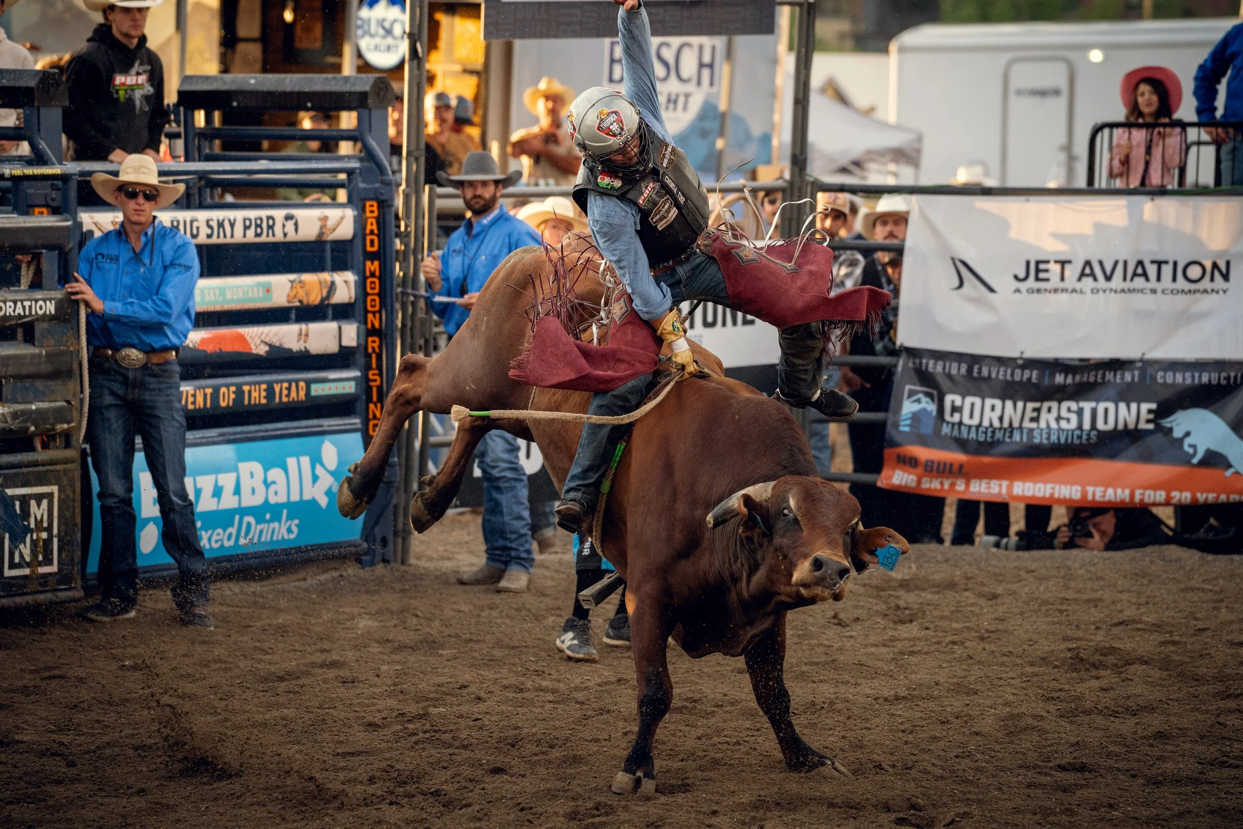 A cowboy riding a bucking bull during a rodeo event, with spectators and event staff in the background.