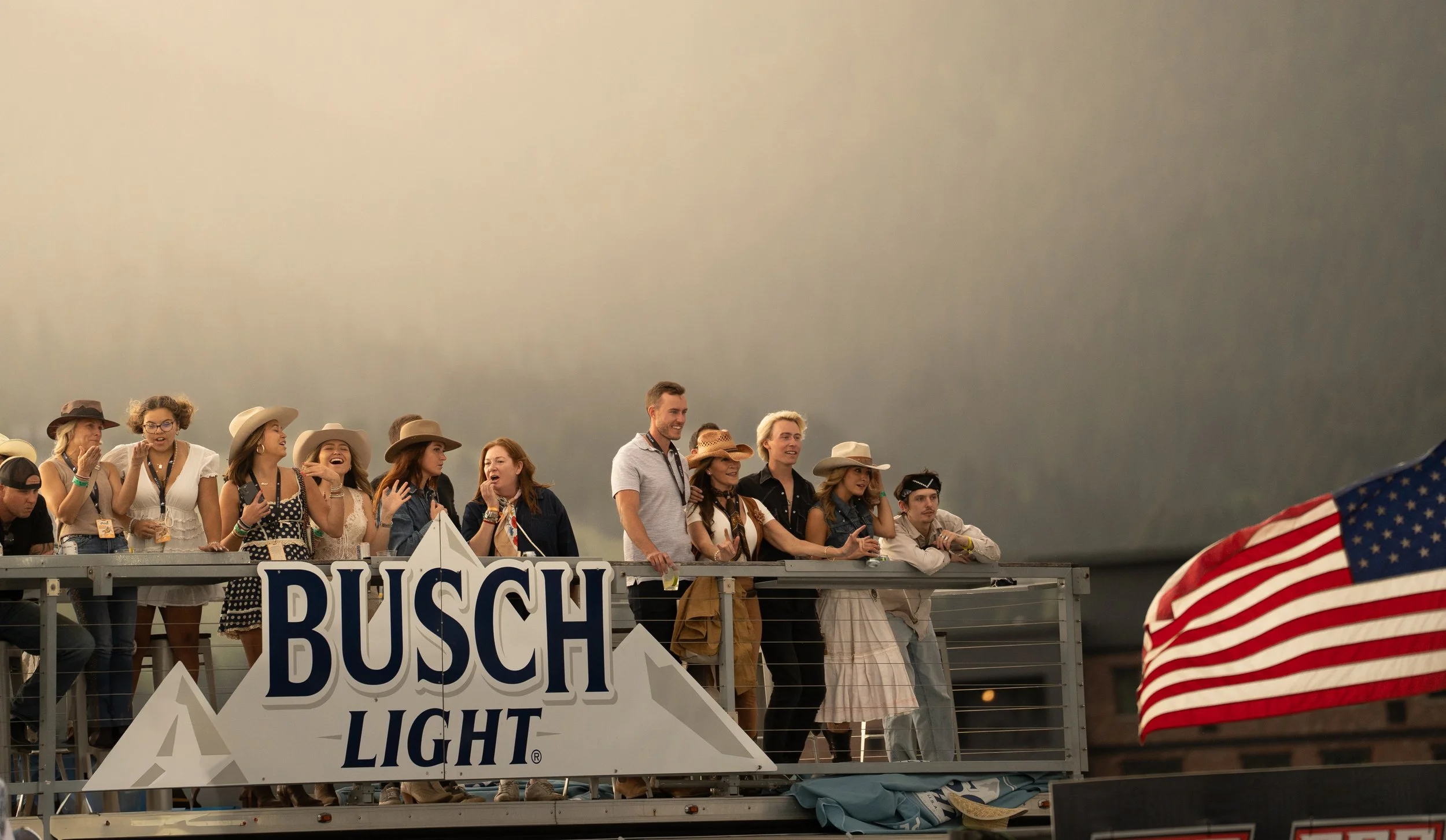 Group of people wearing cowboy hats standing on a raised platform with a Busch Light sign, American flag in the foreground, under gray sky.
