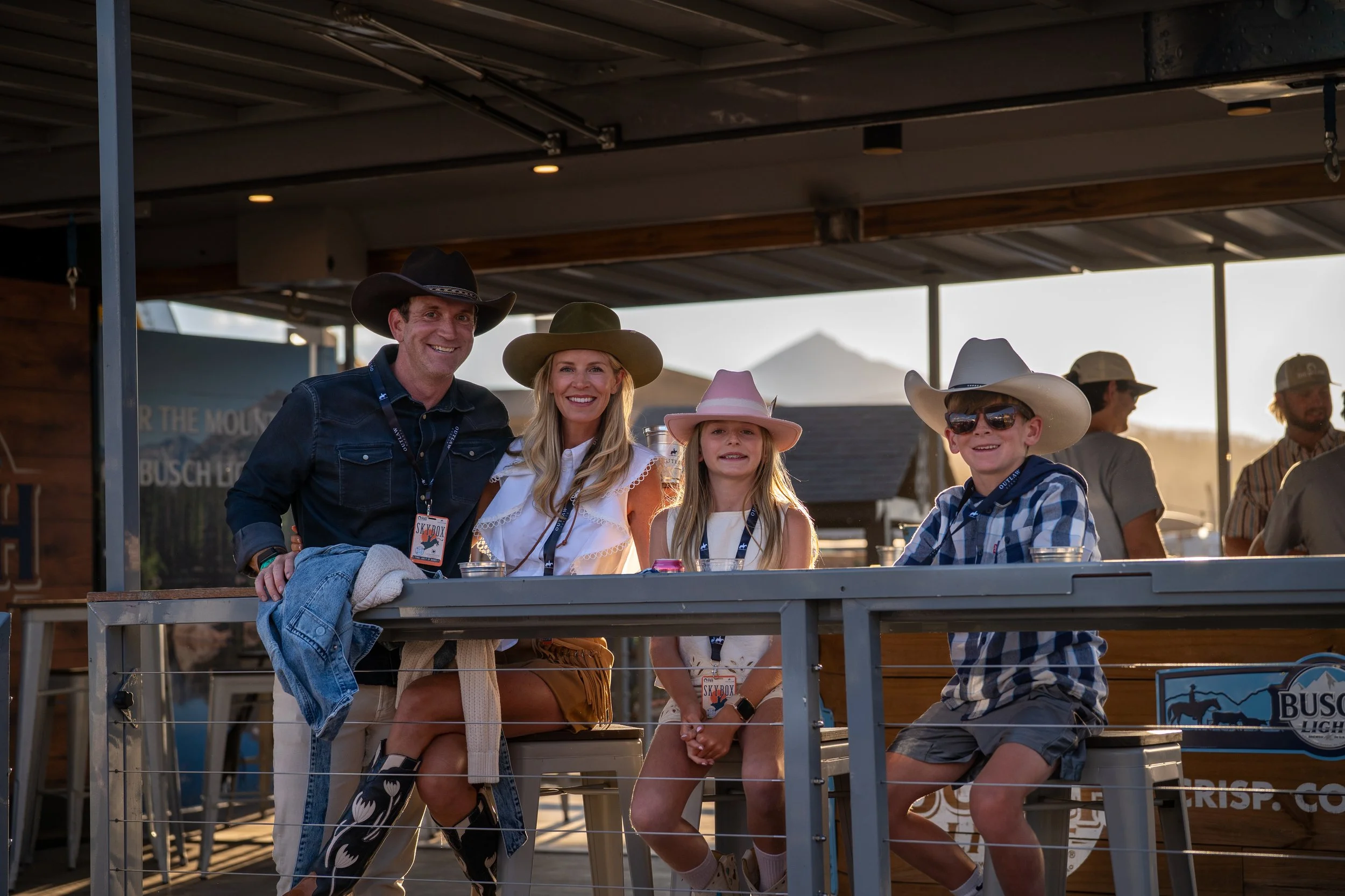Family at an outdoor event wearing cowboy hats, including two adults and two children, with a bar or food stand in the background.