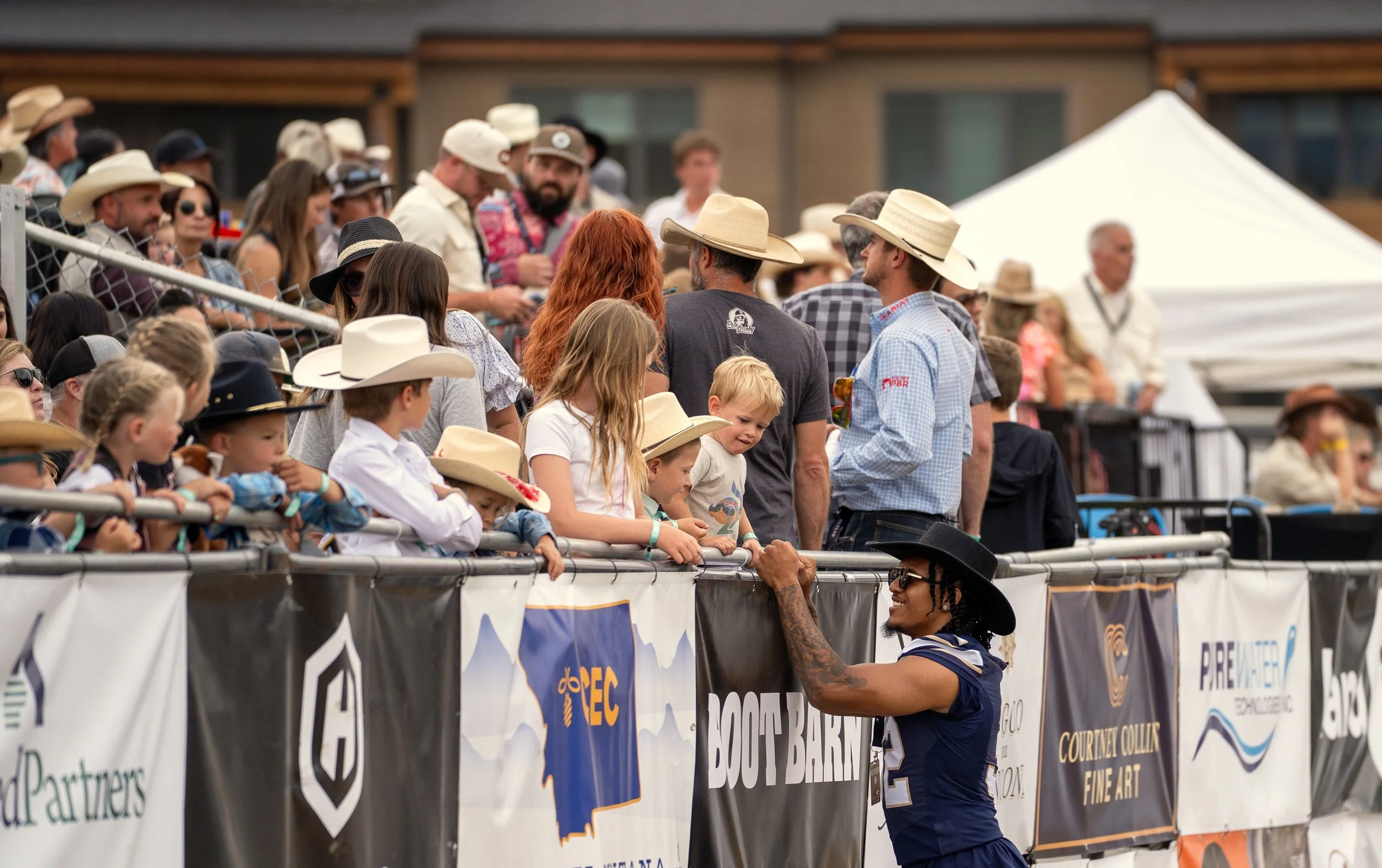 A woman with long braided hair in a cowboy hat and sunglasses interacts with a crowd of children and adults at an outdoor event, with many wearing cowboy hats, some leaning on a barrier.