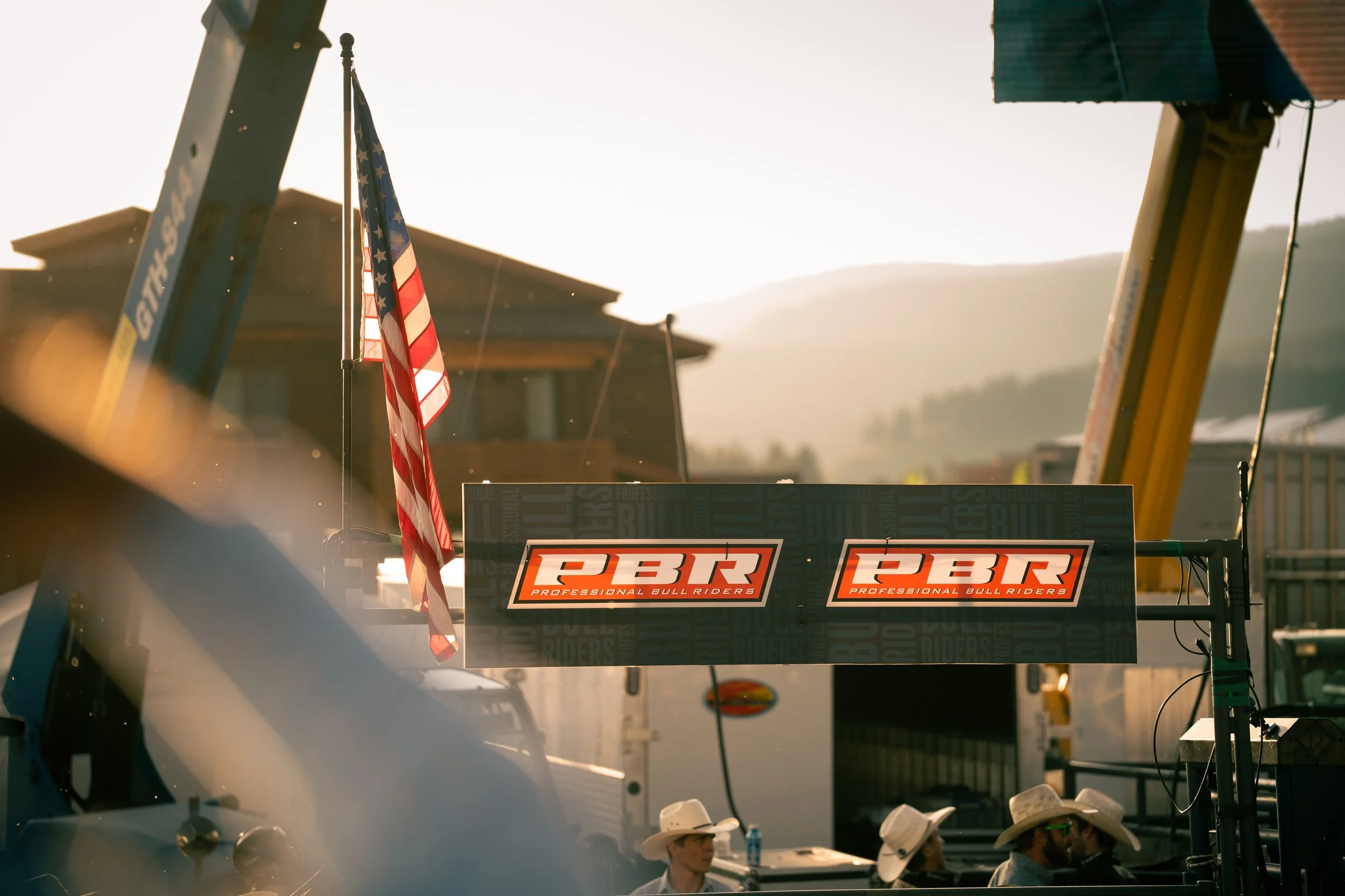 American flag, two signs with 'GBR Professional Bull Riders' logo, in outdoor rodeo or bull riding event setting with people wearing cowboy hats.