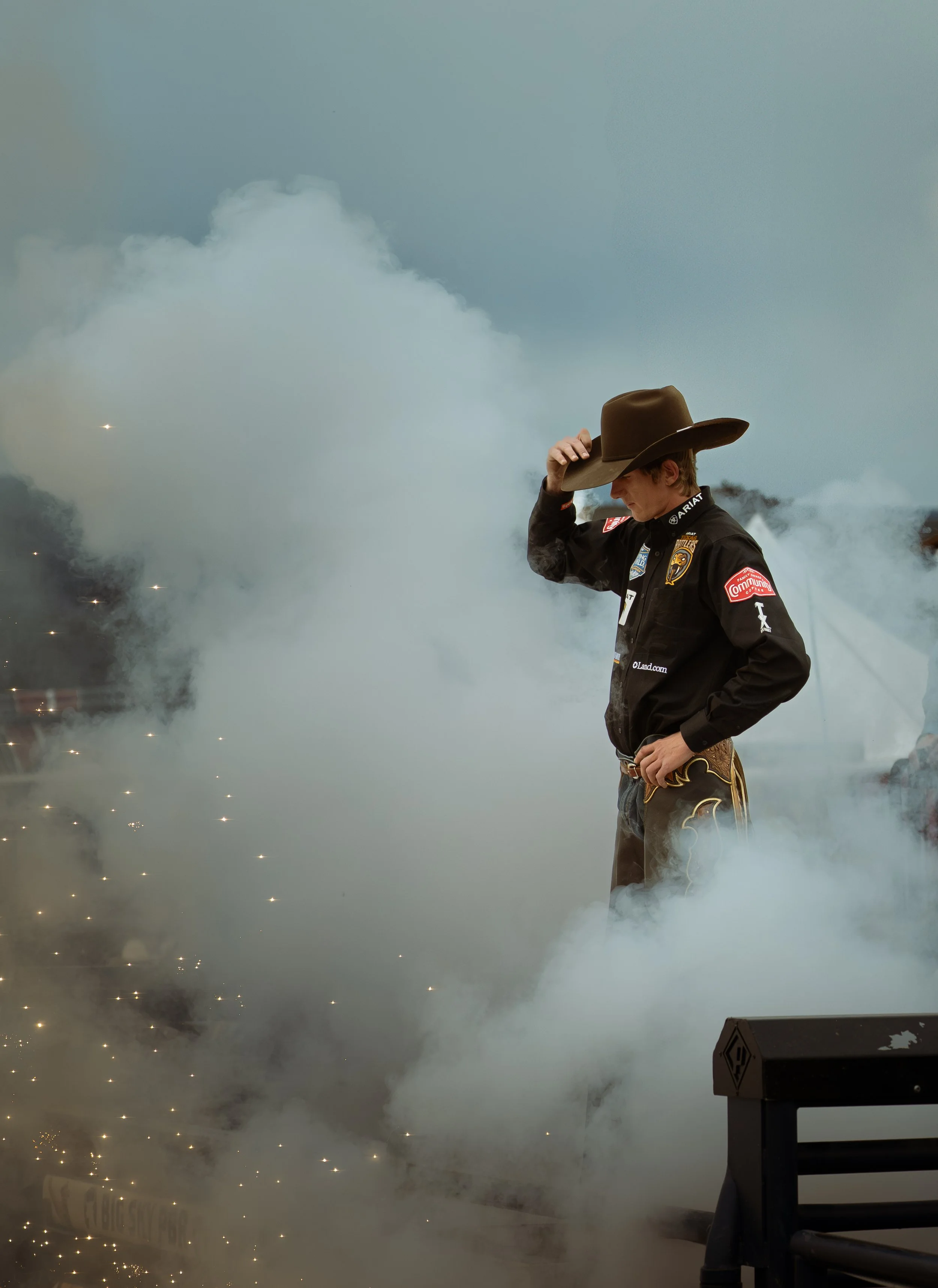 A cowboy dressed in a black jacket and wearing a cowboy hat stands amidst smoke and sparks, with a cloudy sky in the background. Used as the cover photo for the 15th issue of the Explore Big Sky newspaper
