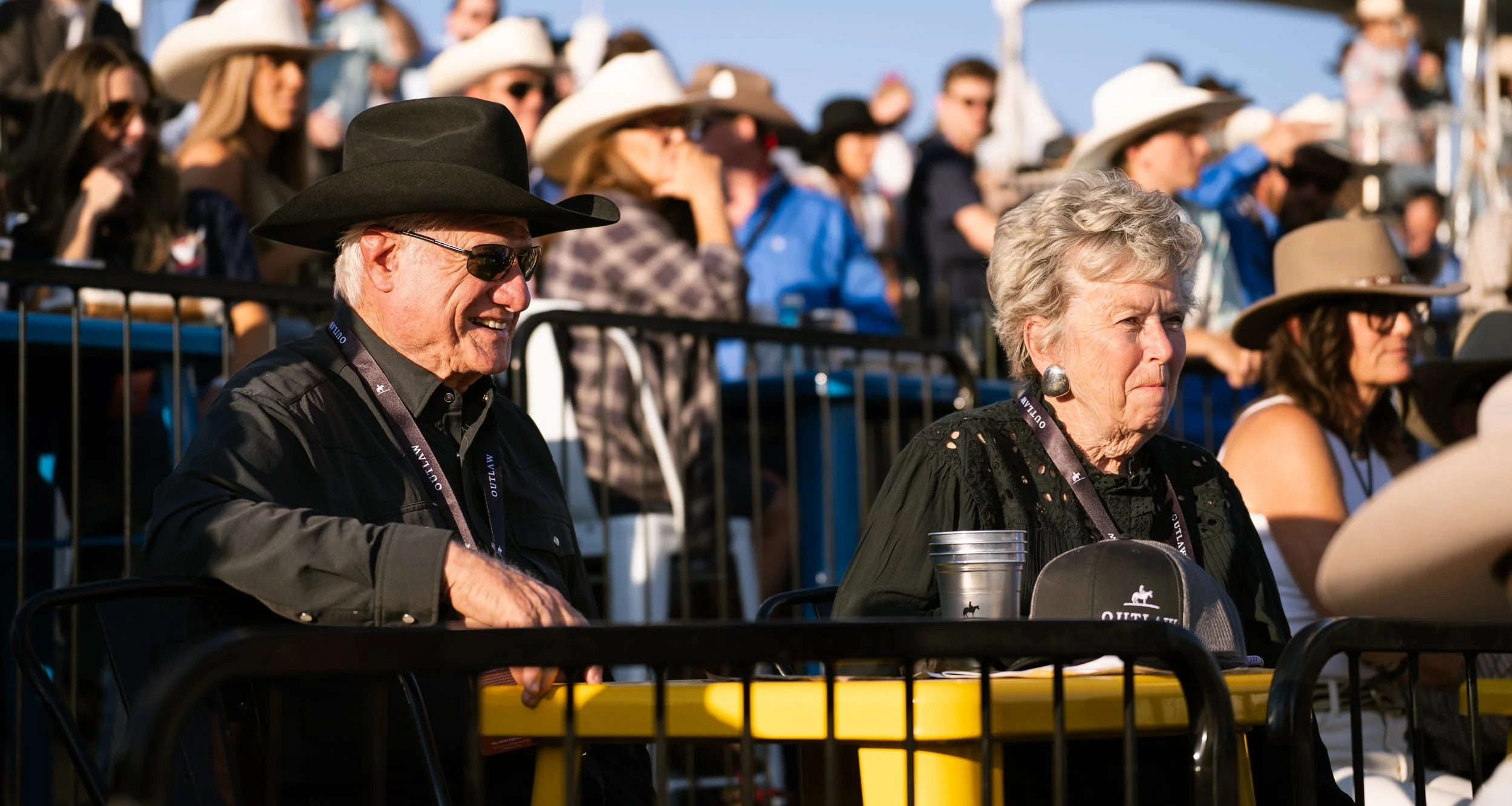 Crowd of people at an outdoor event, including an elderly man wearing a black cowboy hat, sunglasses, and black shirt, and an elderly woman with short gray hair, wearing a black top and earrings, sitting at a yellow table.