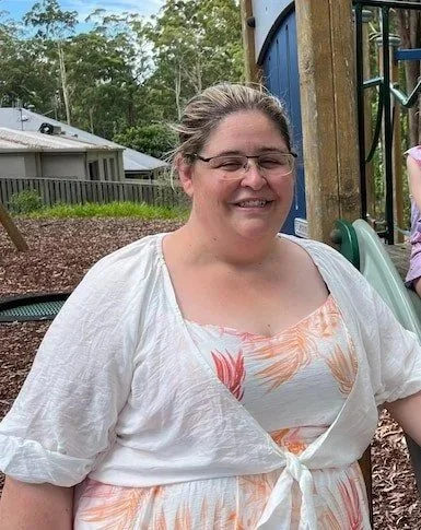 A woman and two children smiling on a playground slide, surrounded by trees.