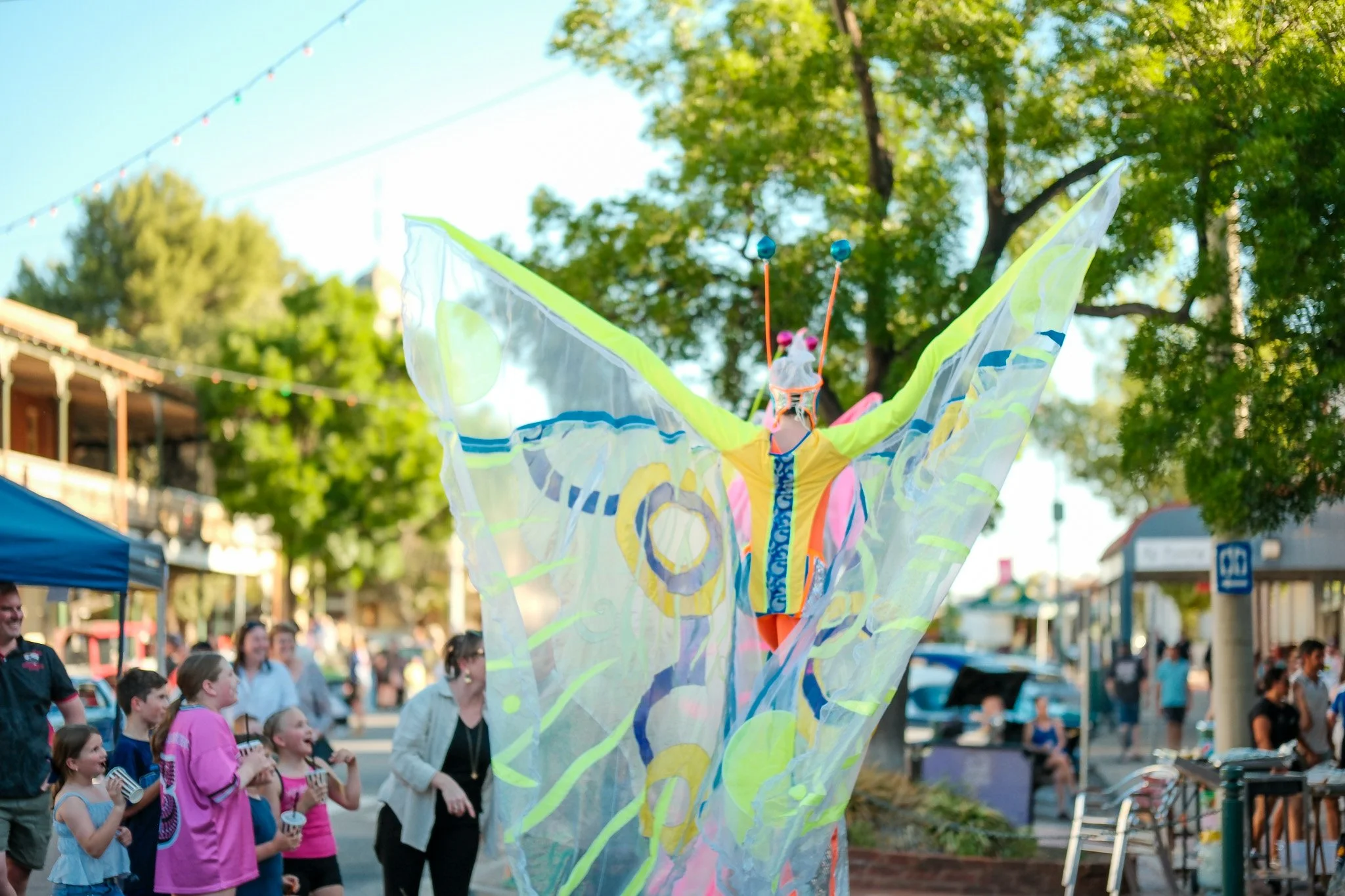 A person on stilts wearing colourful butterfly wings during the Narrandera Spring Fair, with a crowd of children and adults watching and enjoying the event.