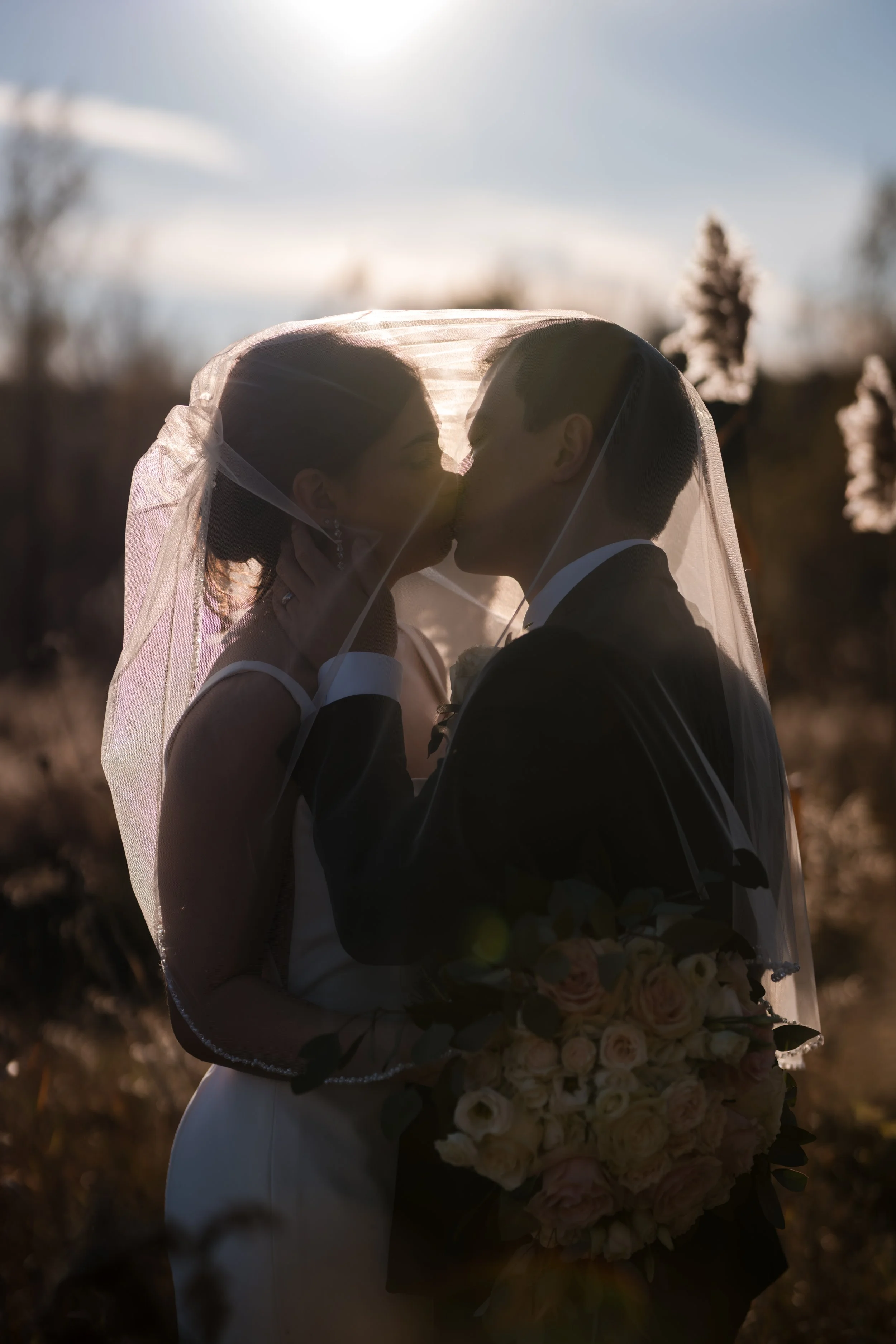 A bride and groom sharing a kiss under a veil during an outdoor wedding at sunset.