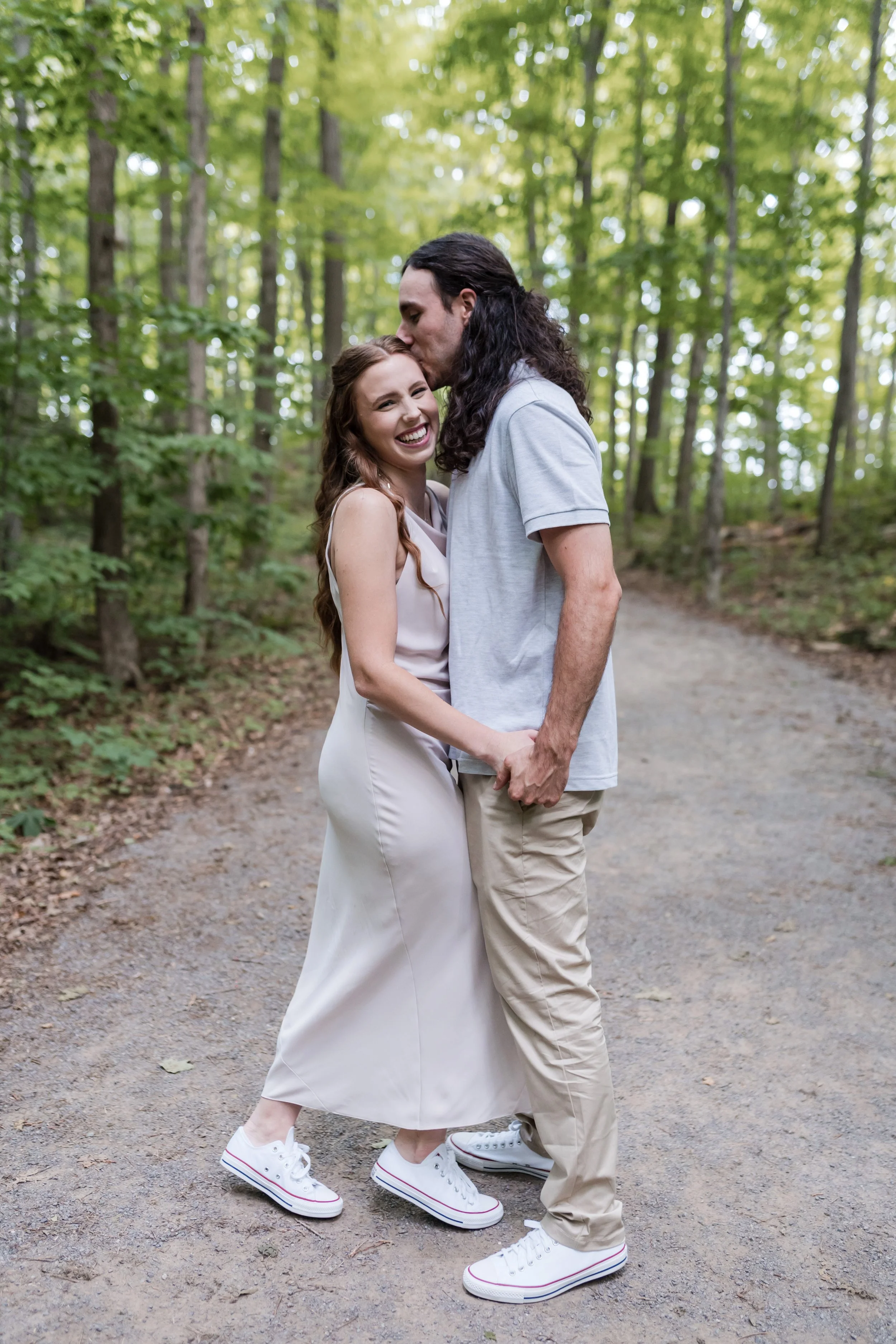 A couple holding hands and smiling in a forest with tall trees and green leaves. The man is kissing the woman's forehead, and she is smiling happily. They are wearing casual clothes and white sneakers.