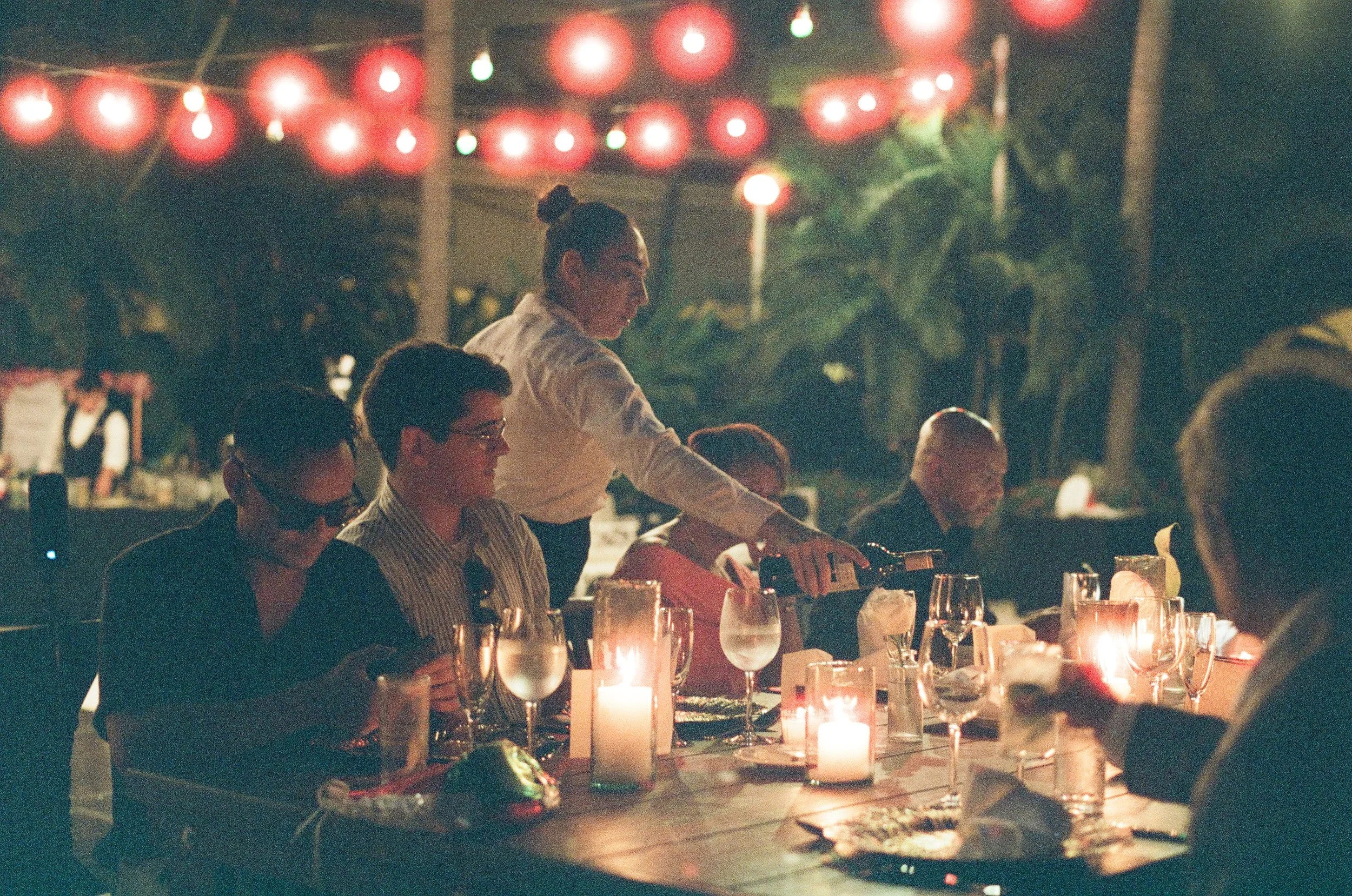 A woman serving drinks at a dinner table decorated with lit candles, with red lanterns hanging overhead during an outdoor evening gathering.