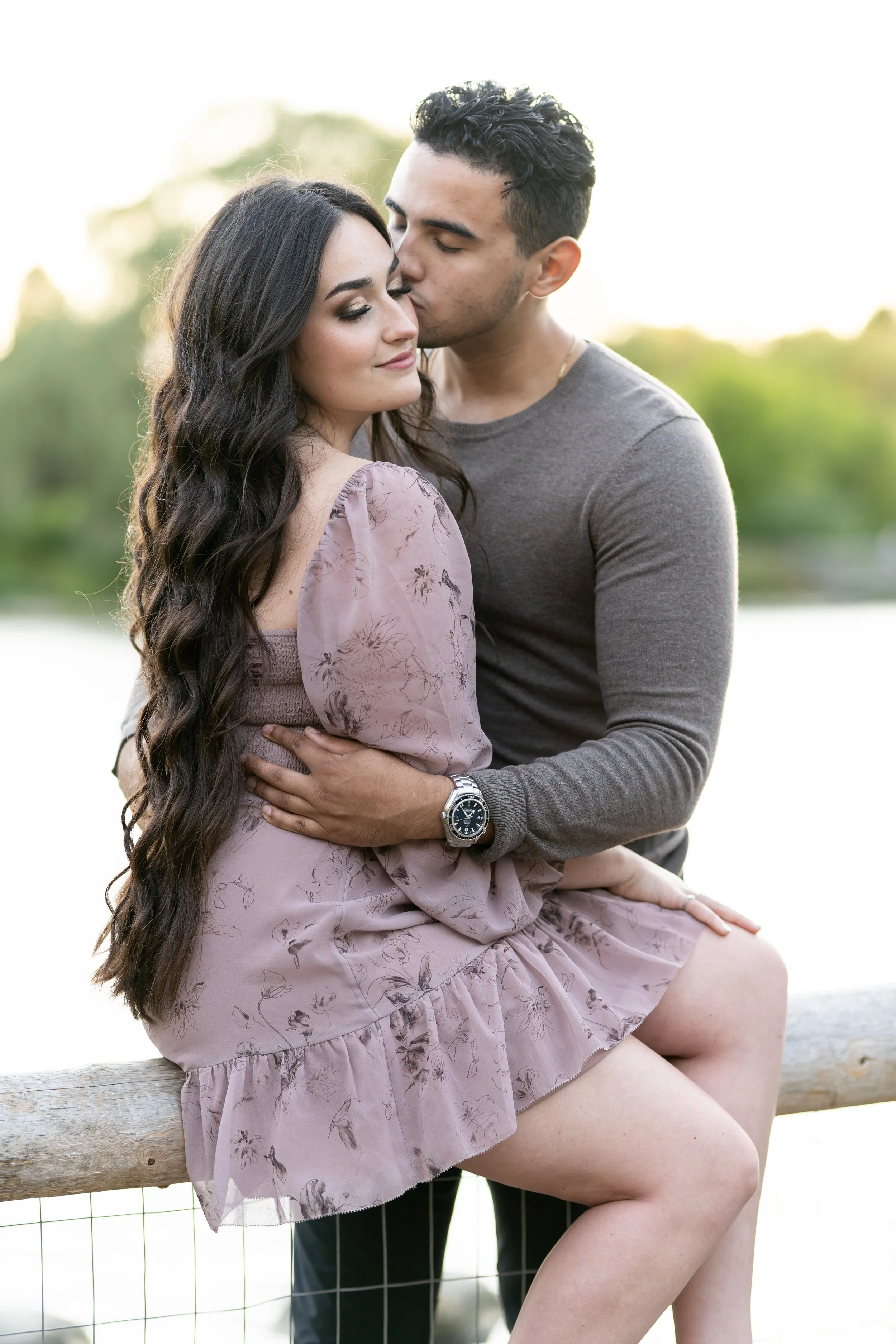 A young couple sitting outdoors on a wooden bench near a body of water. The man is kissing the woman's forehead, and she has a content expression with her eyes closed. The background is blurred greenery with sunlight.
