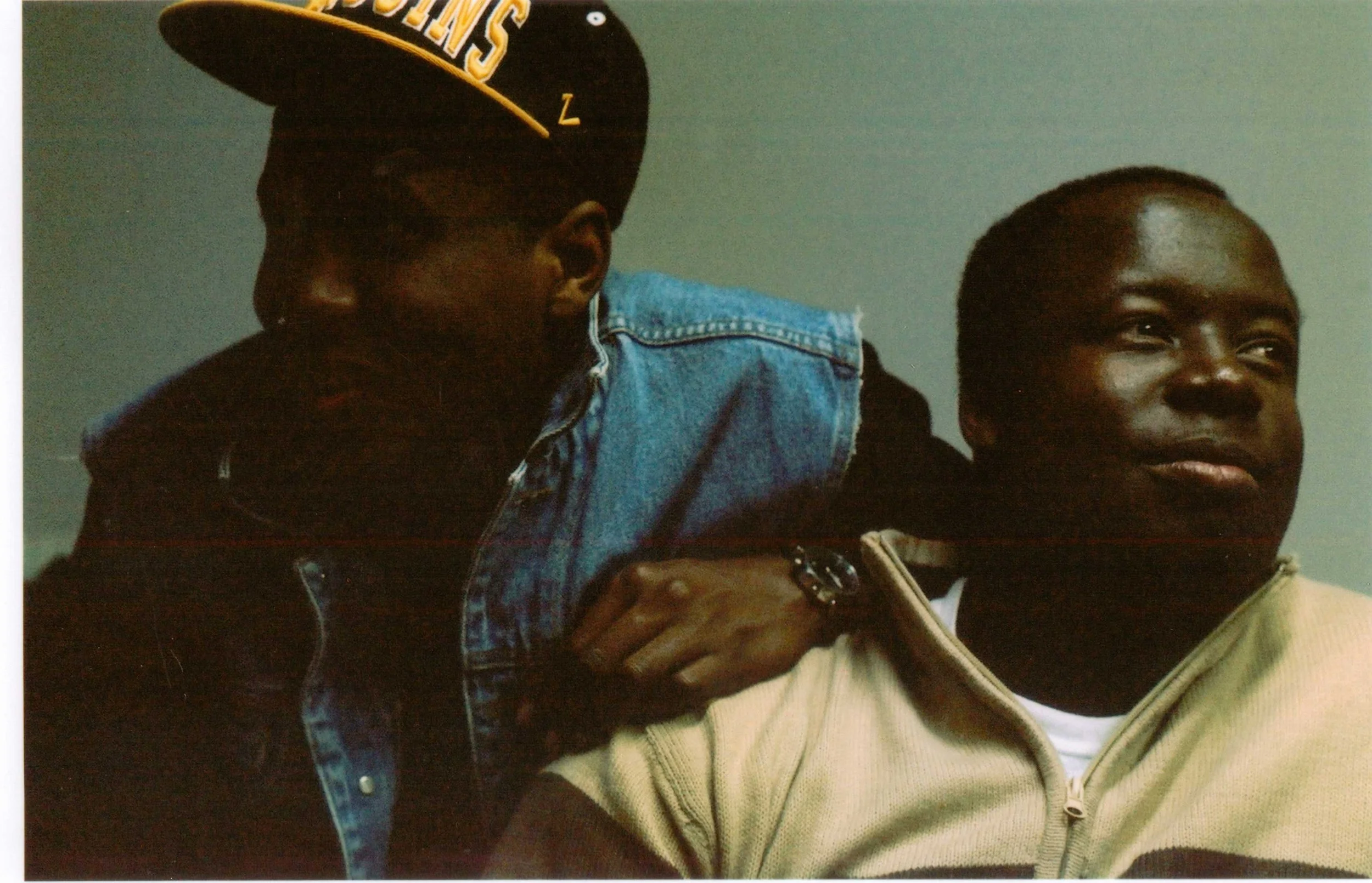 Two young men are close together, one in a yellow and black jacket, the other in a beige hoodie, both with dark skin and short hair. The man in the jacket is looking at the other, who is smiling slightly. The background is plain with a neutral color.