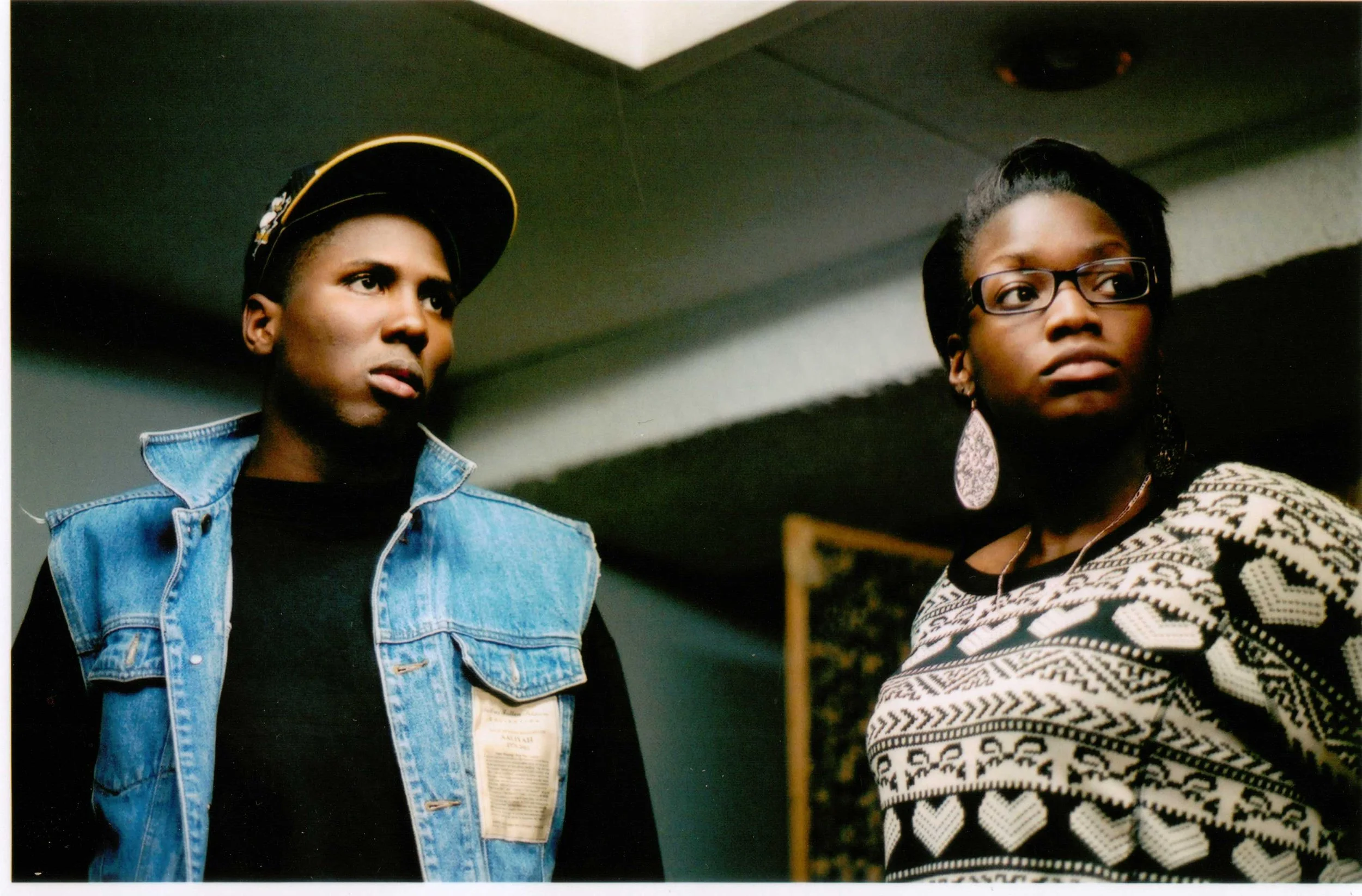 Two women standing indoors, one wearing a black cap with yellow and white details, a denim jacket, and a black top, and the other wearing glasses, large earrings, and a patterned sweater.