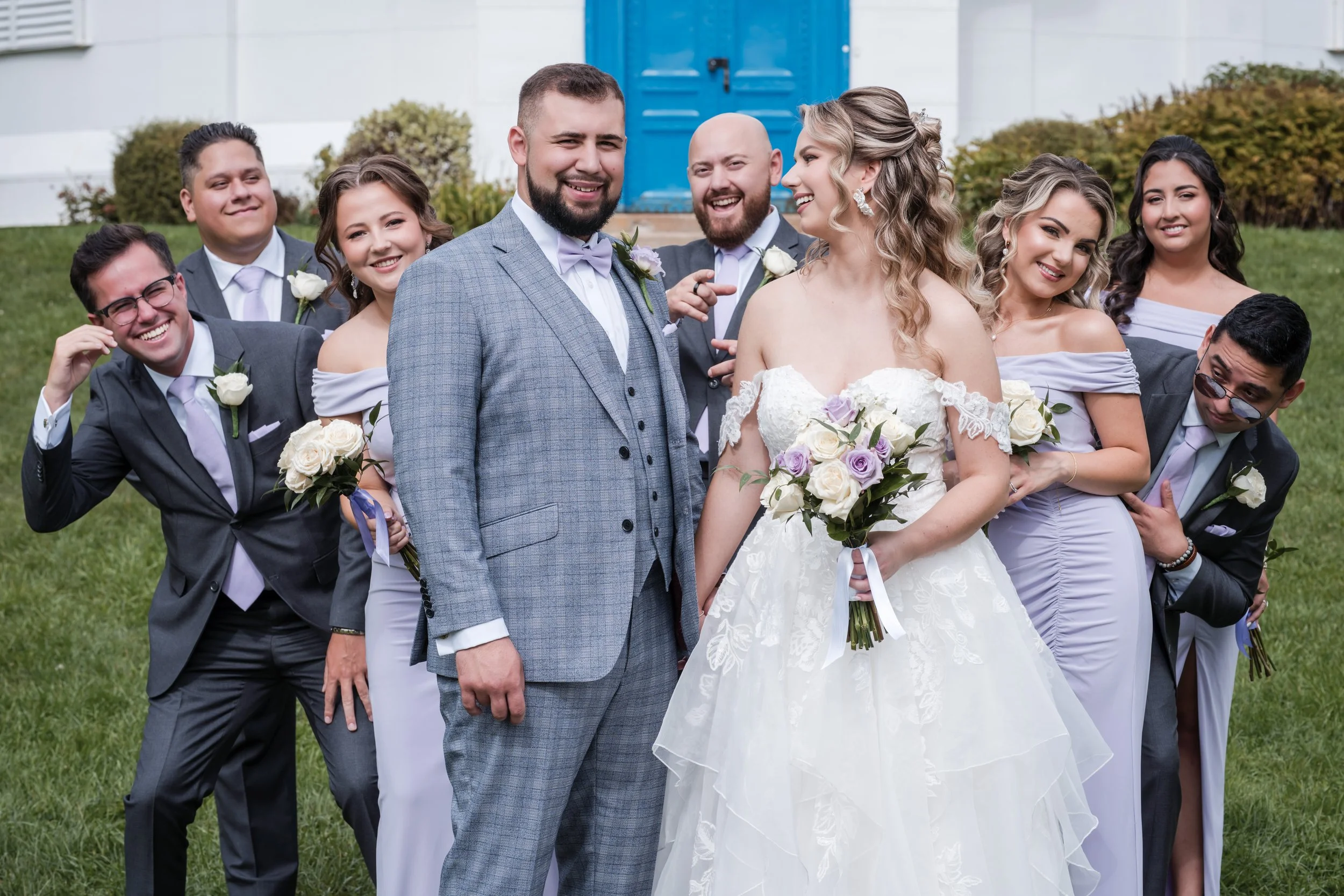 A group of people dressed in formal attire, including a bride with a bouquet, standing outdoors on a lawn, smiling and posing for a photo.