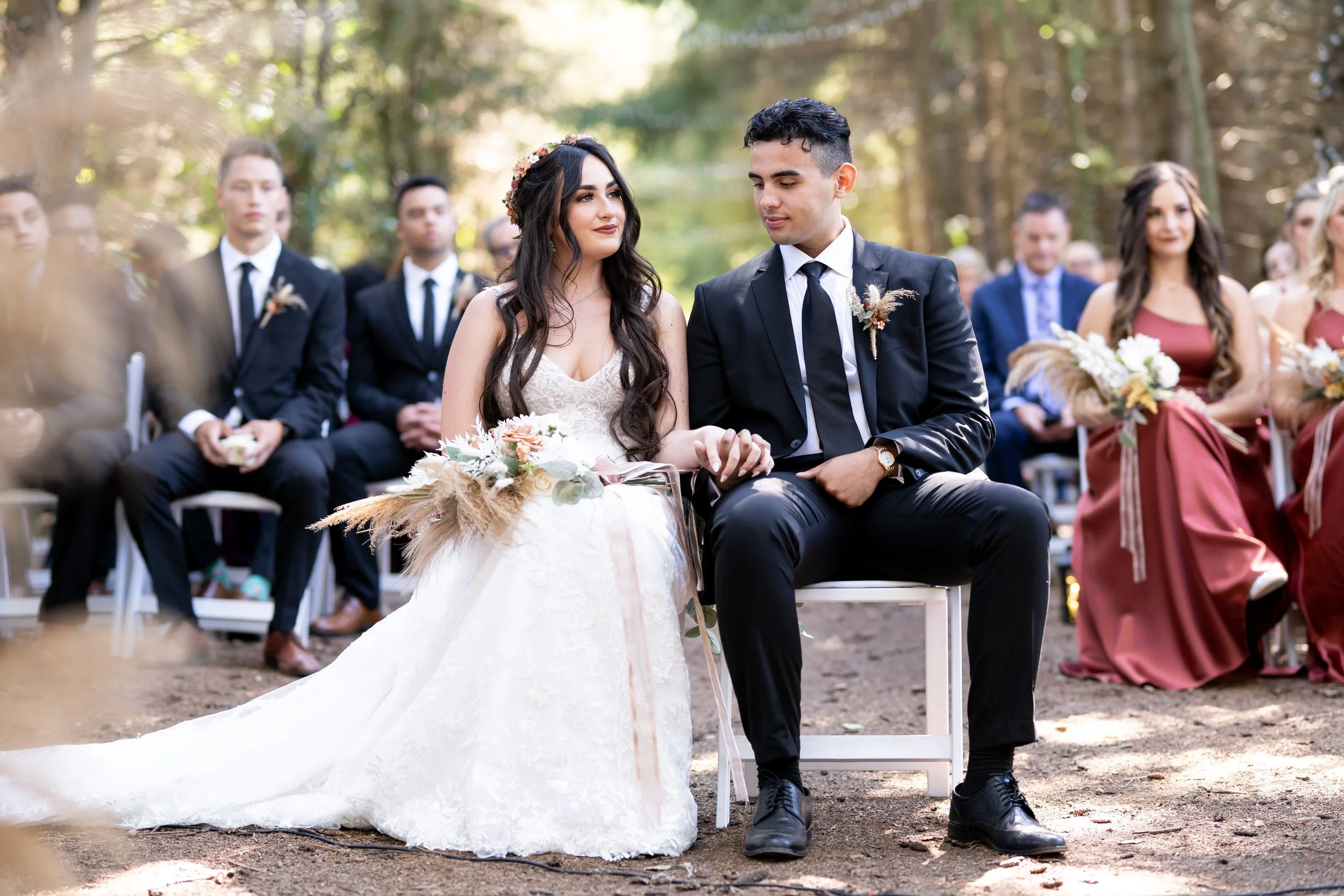 A bride and groom sitting next to each other during an outdoor wedding ceremony in a wooded area, surrounded by friends and family.