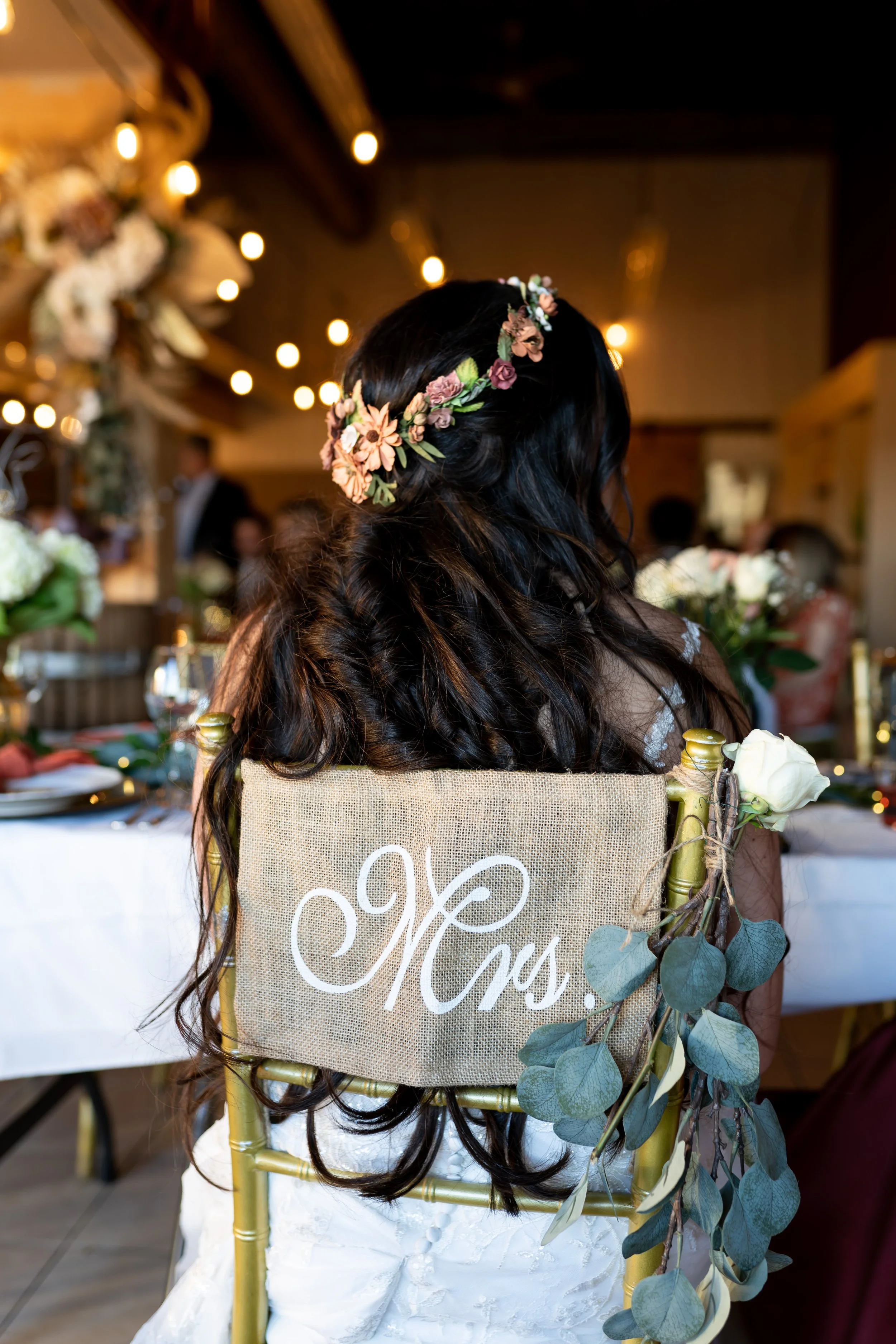 Back of a woman with long dark hair wearing a floral crown, seated at a decorated table with lights and floral arrangements, in a warmly lit indoor event setting.