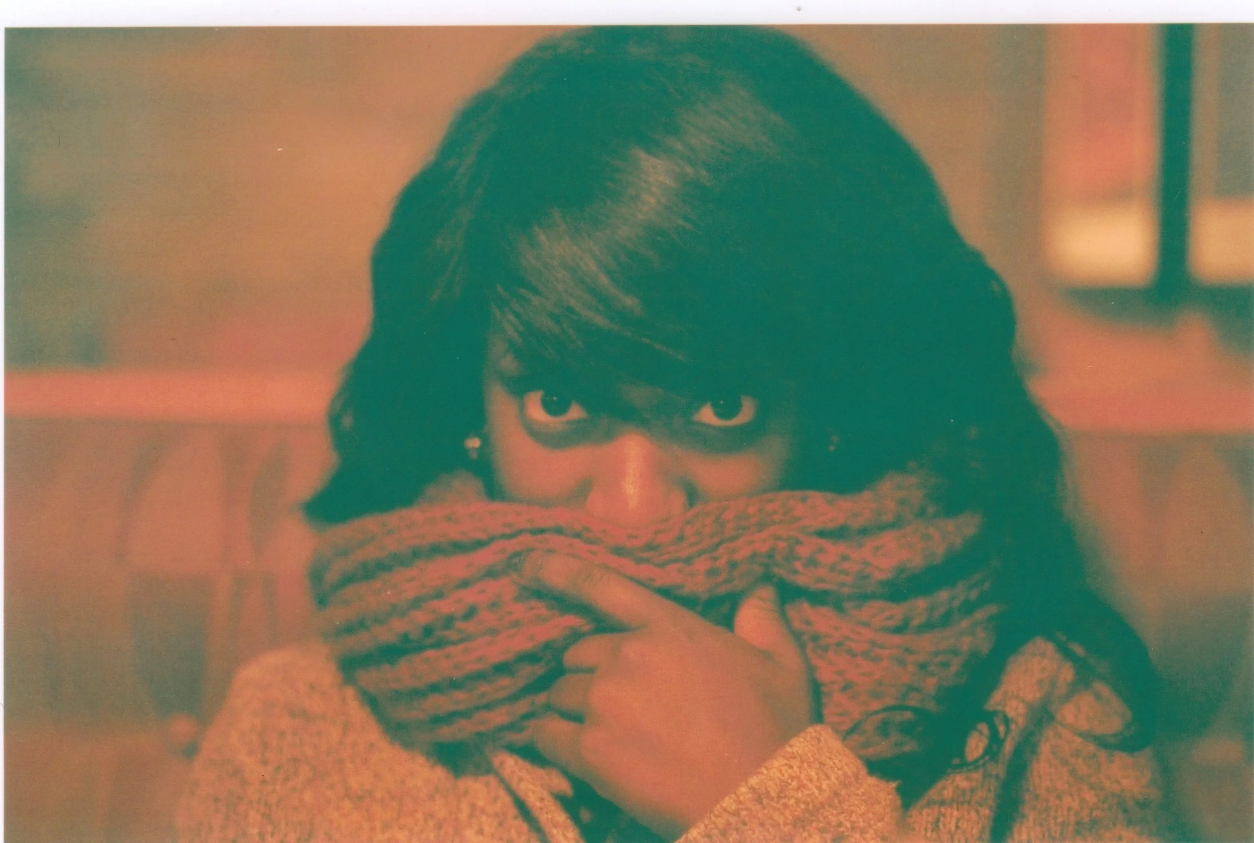 Close-up of a person's face, with dark hair covering part of their forehead, partially hidden behind a knitted scarf or blanket, and wearing glasses.