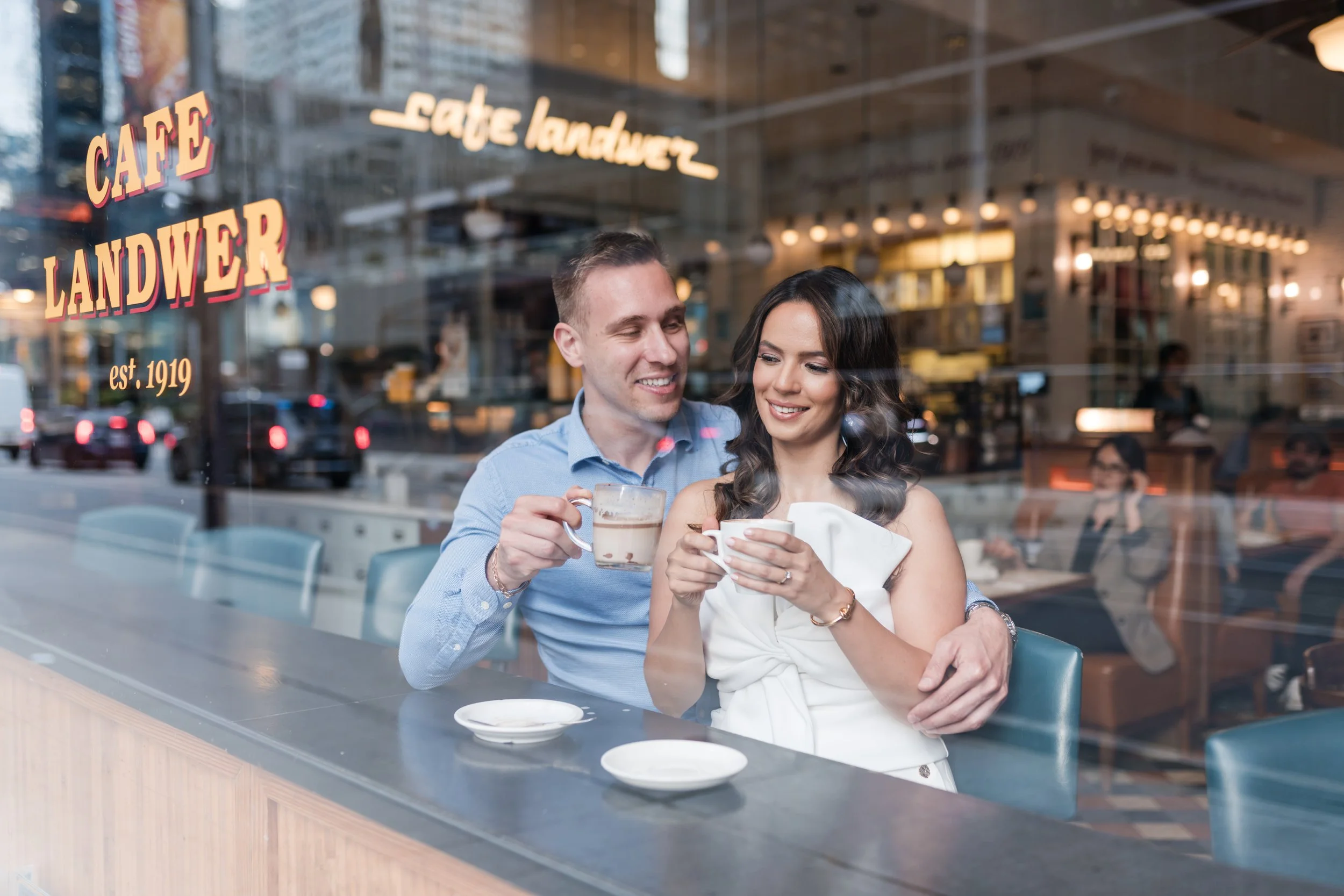 A man and woman sitting inside a café, smiling and holding coffee mugs, seen through the café window.