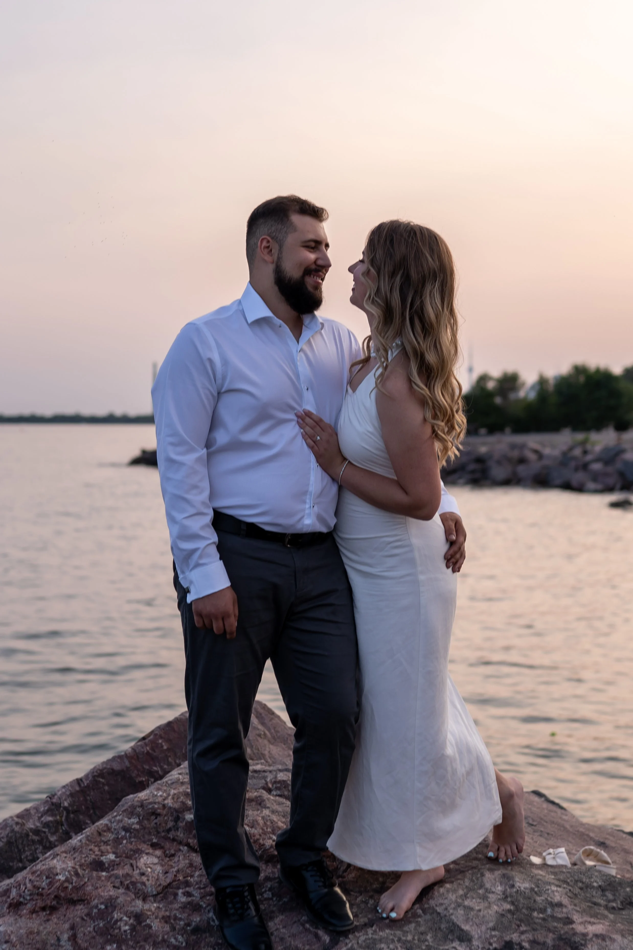 A couple sharing a romantic moment by a body of water during sunset. The man is wearing a white dress shirt and dark pants, while the woman is in a white dress. They are standing on rocks, facing each other closely, with the man's arm around the woma
