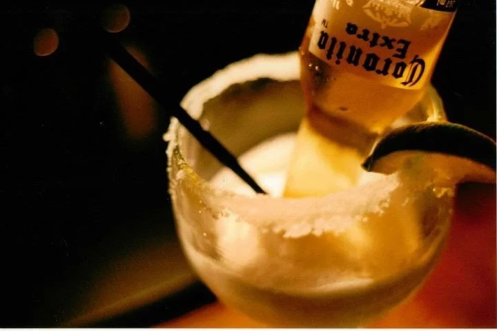 A margarita glass with salt on the rim, containing a Corona beer bottle floating inside and a lime wedge on the edge, placed on a dark surface.