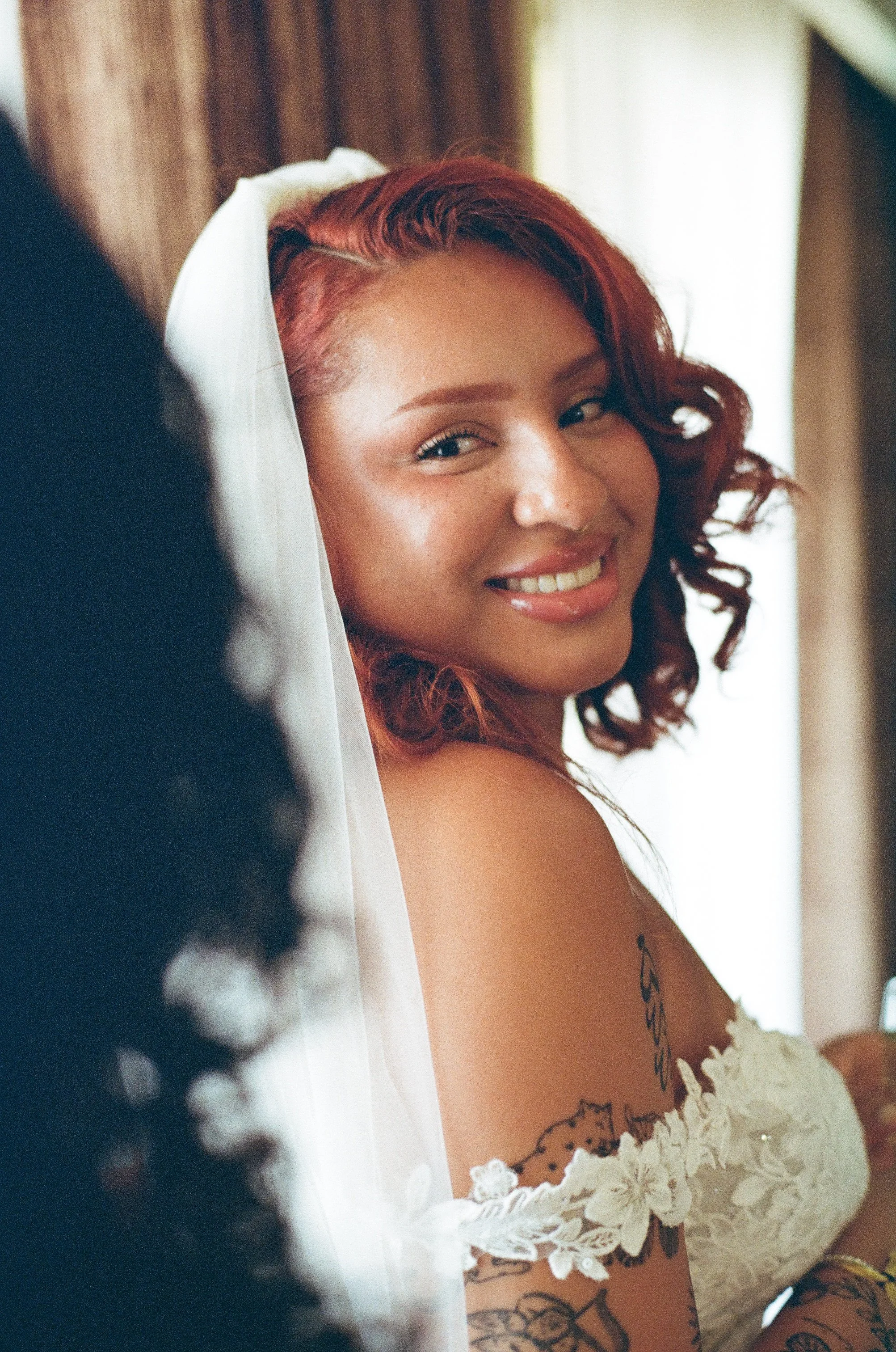 A woman with red curly hair and tattoos smiling with white lace clothing.