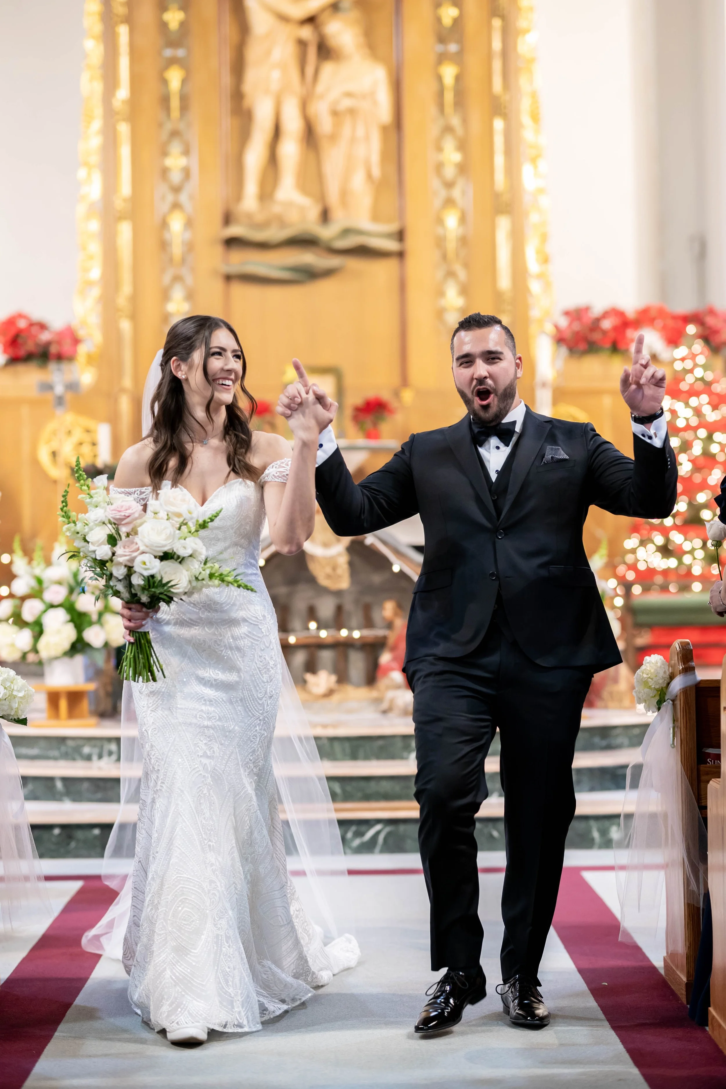 A newlywed couple walking down the aisle inside a church decorated with flowers and Christmas lights. The bride is holding a bouquet of white and pink flowers, while the groom is wearing a black tuxedo and smiling.