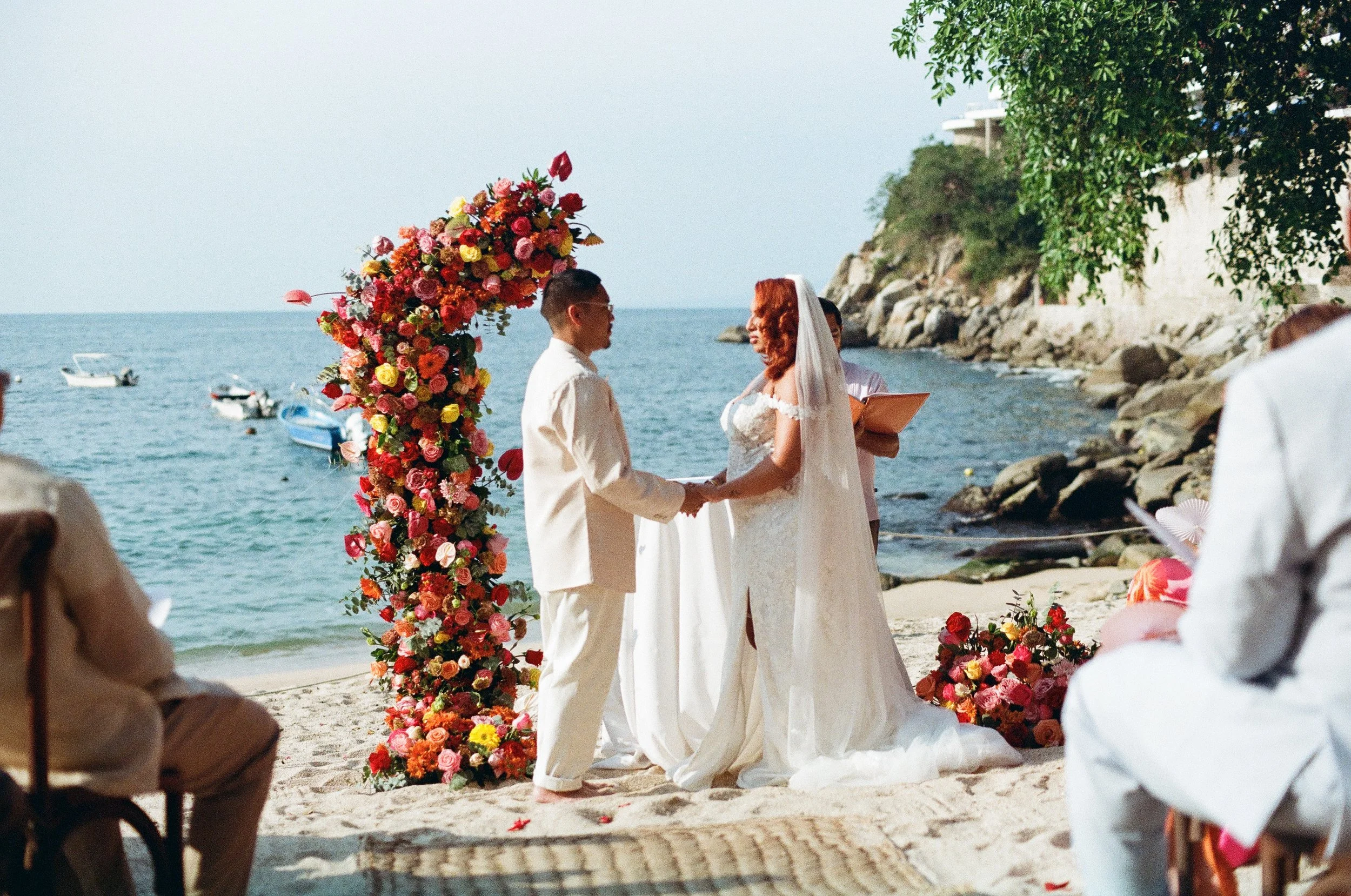 A beach wedding ceremony with a couple holding hands and facing each other, surrounded by vibrant floral arrangements, overlooking the ocean with boats in the water and a rocky shoreline in the background.