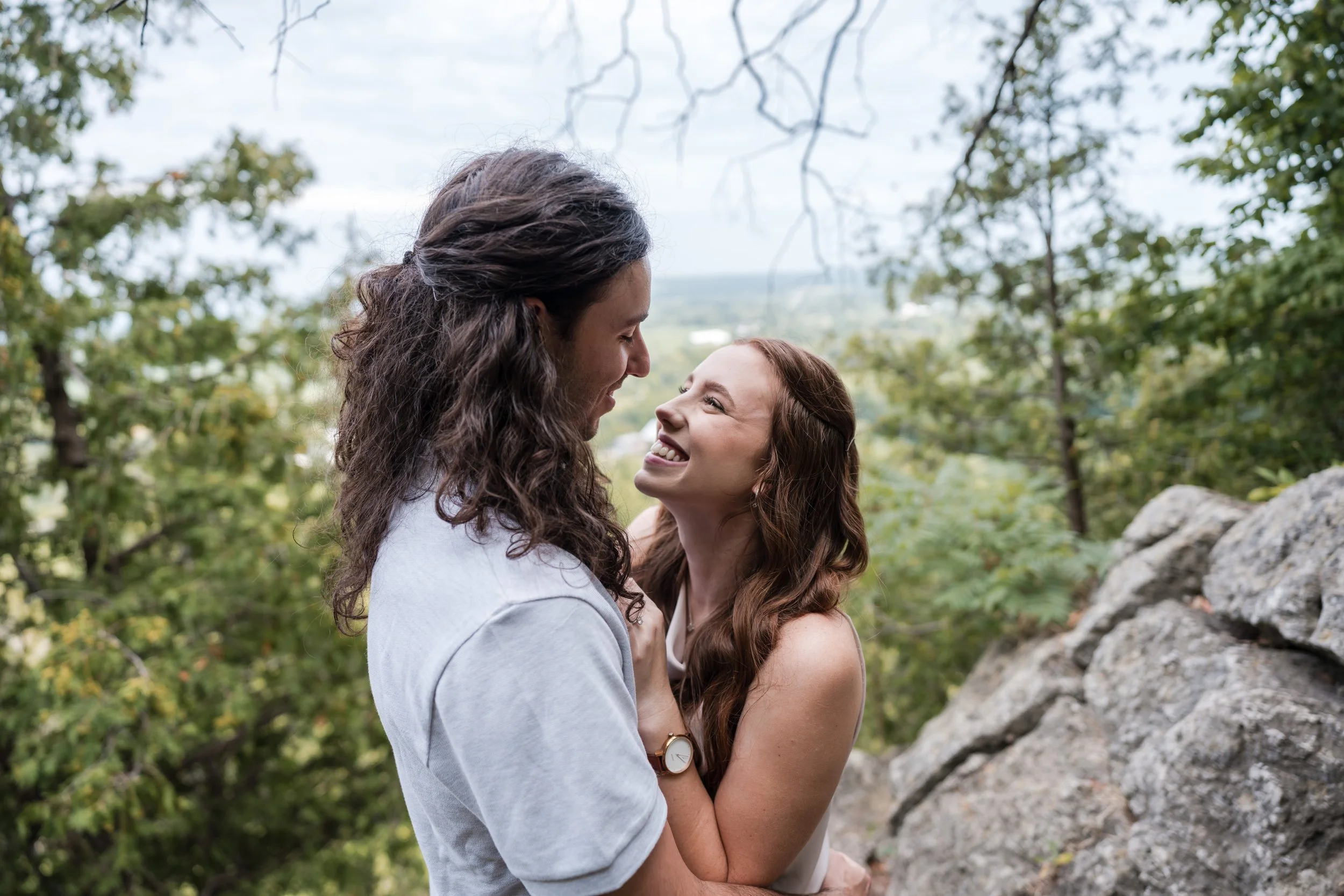 A man and woman embrace outdoors, smiling and looking at each other, with trees and rocks in the background.