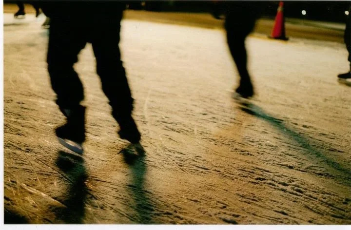 People ice skating outdoors at night on a rink marked by orange cones, with a textured ice surface and shadows cast on it.